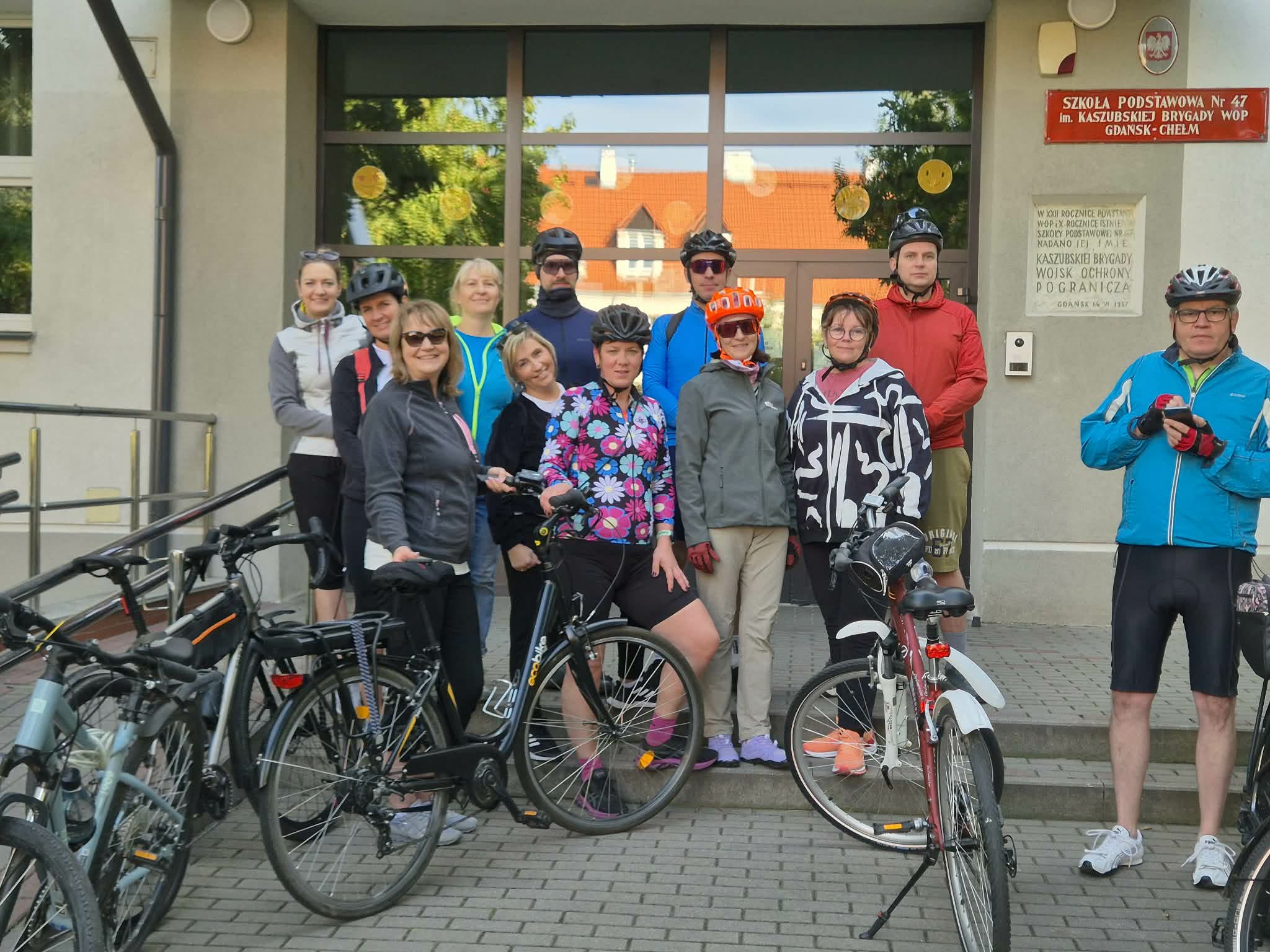 1. Grupa nauczycieli i pracowników szkoły z rowerami po wspólnej przejażdżce. Uczestnicy stoją przed wejściem do Szkoły Podstawowej nr 47 im. Kaszubskiej Brygady WOP w Gdańsku-Chełmie. Na pierwszym planie widoczne są rowery, a na ścianie budynku znajduje się tablica z nazwą szkoły.