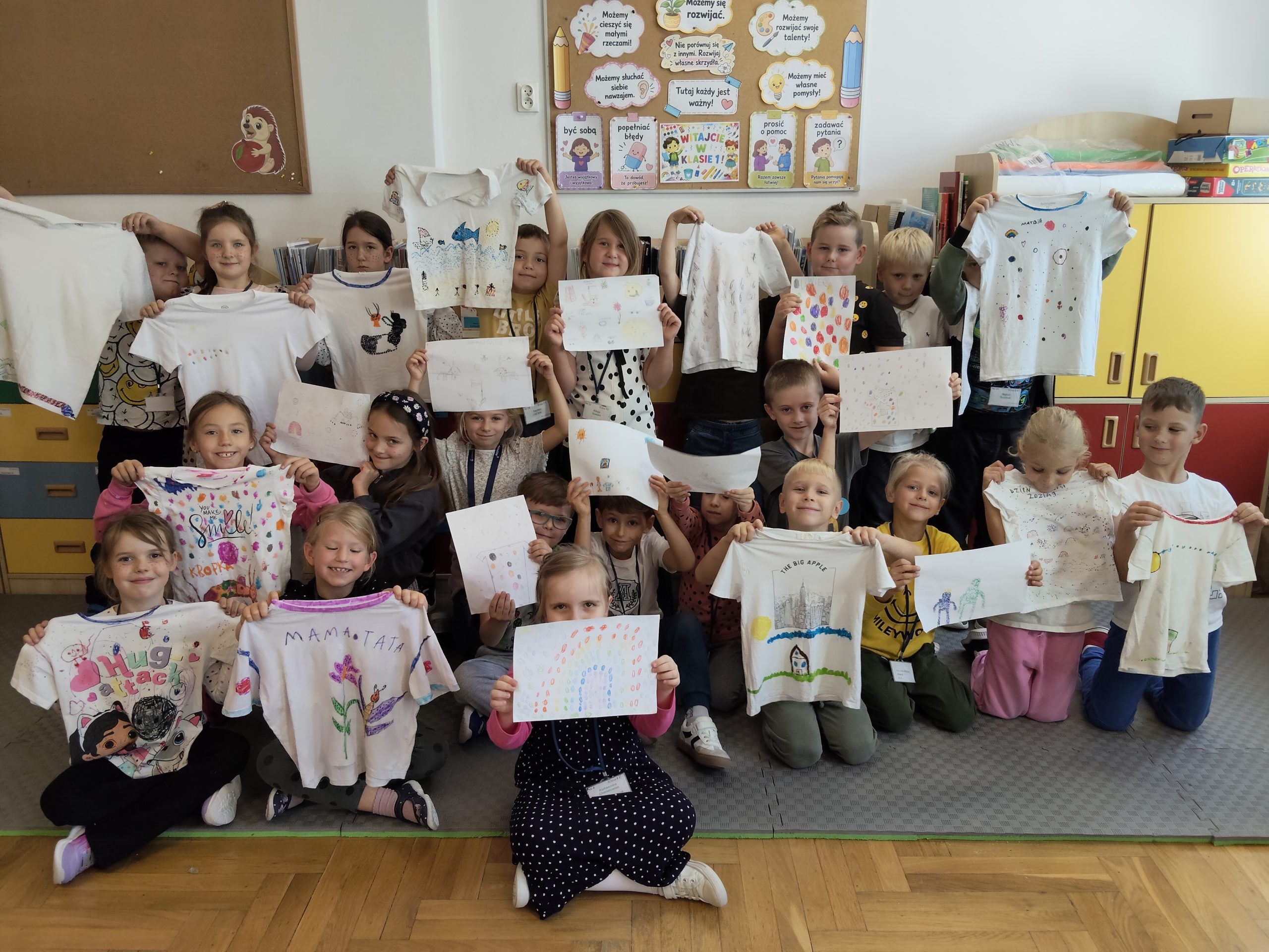 Dzieci w sali lekcyjnej świętują Dzień Kropki, prezentując ozdobione przez siebie koszulki i rysunki.