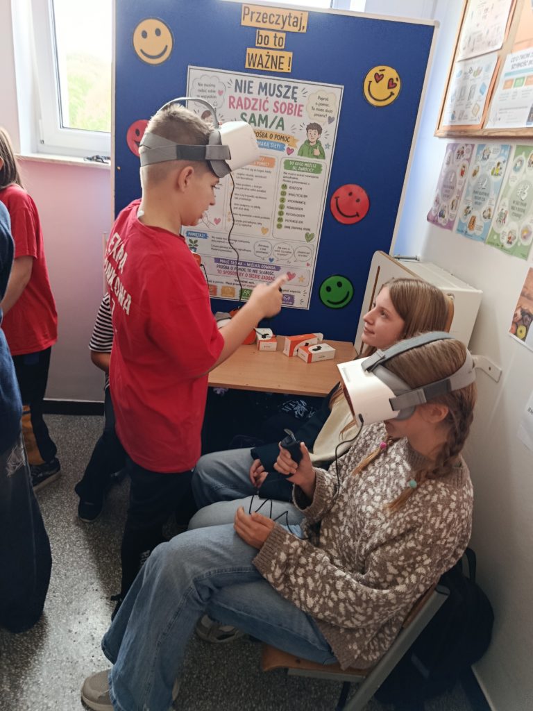 Na pierwszym planie uczeń Koła PP HOPR i dwie siedzące uczennice z okularami VR. W tle tablica na temat zdrowia psychicznego.