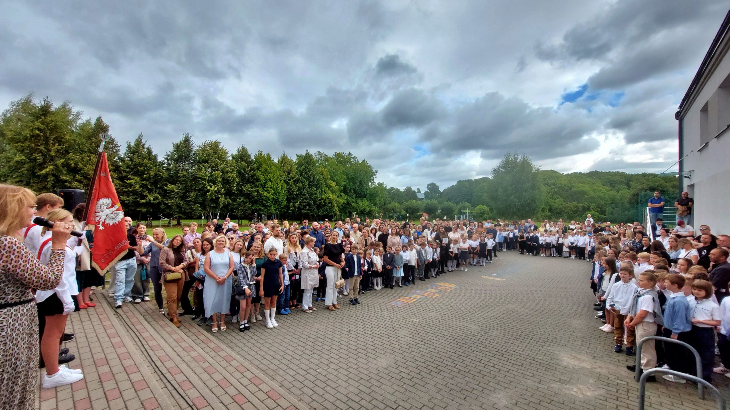 Szeroki kadr na szkolny dziedziniec pełen uczniów, rodziców i nauczycieli podczas uroczystości rozpoczęcia roku szkolnego pod gołym niebem.