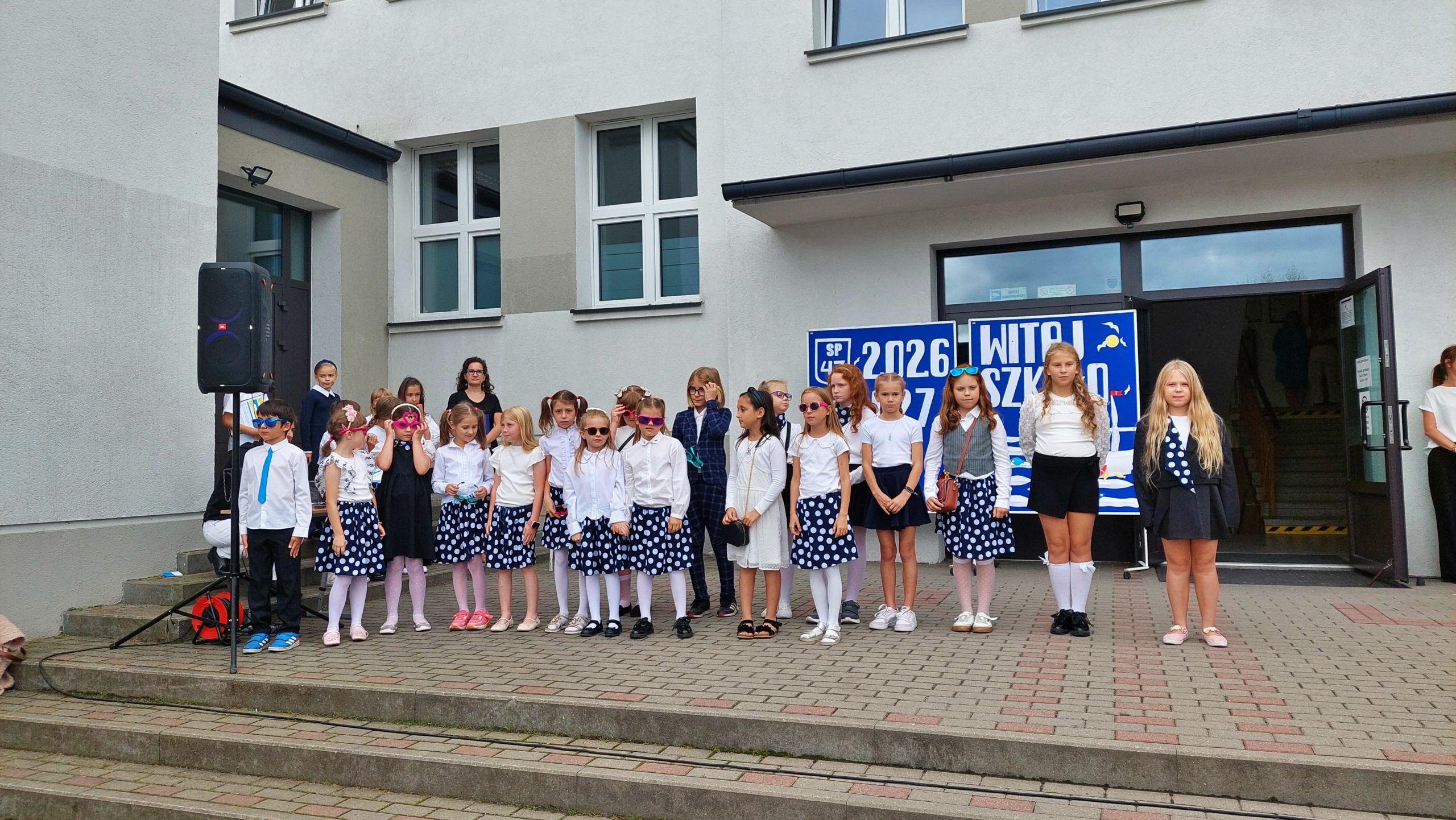 Grupa młodszych uczniów w odświętnych strojach w kropki, stojących na szkolnych schodach podczas apelu inaugurującego rok szkolny.