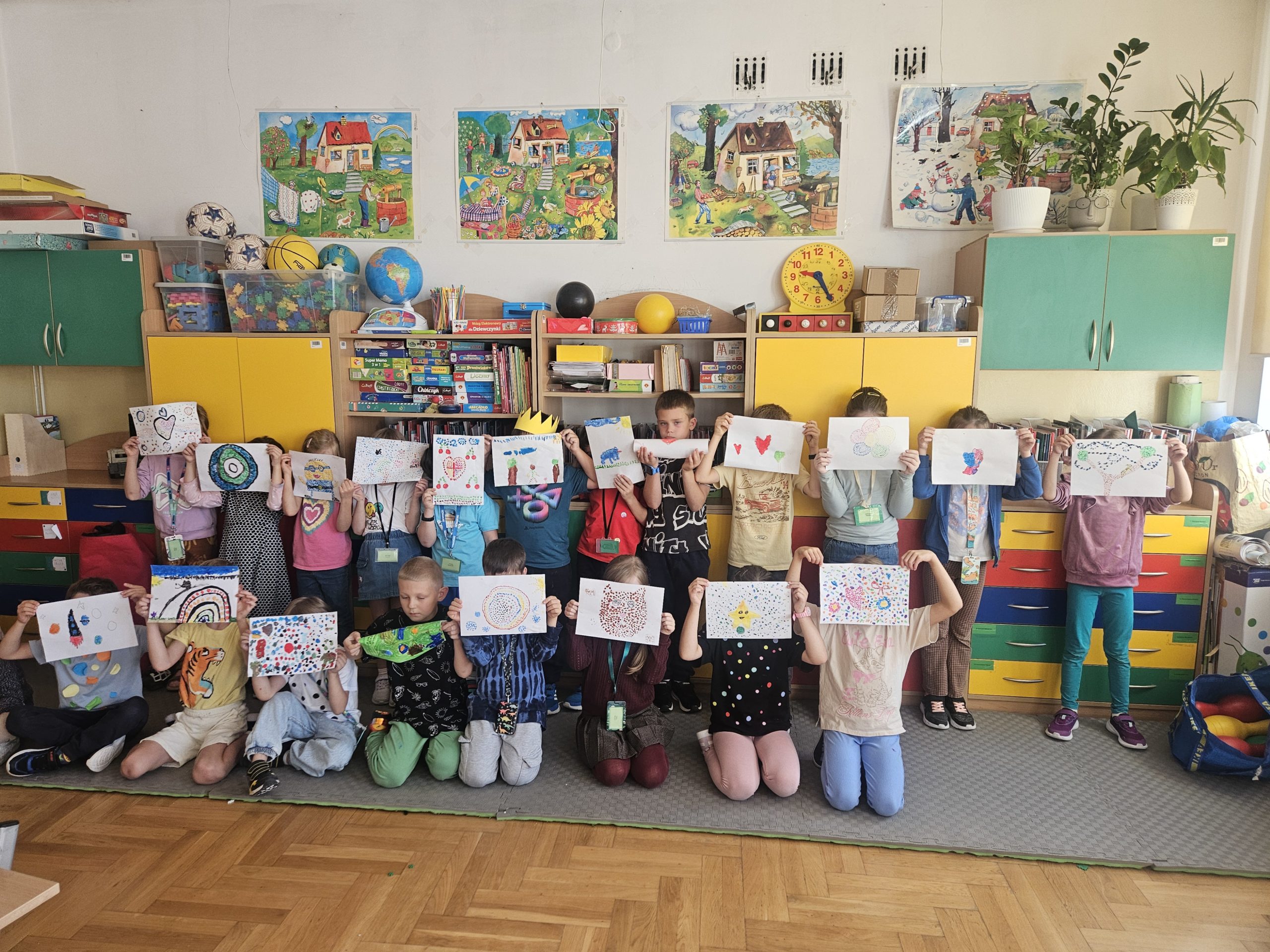 Grupa dzieci w klasie prezentuje swoje prace plastyczne, trzymając je przed twarzami. Na zdjęciu widać kolorowe szafki, półki z zabawkami i materiałami dydaktycznymi, obrazy na ścianie oraz rośliny stojące na szafkach.