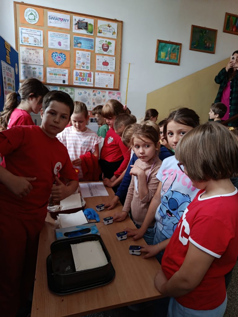 Na pierwszym planie uczeń Koła PP HOPR częstujący ciastem. Obok niego dziewczynki z klas młodszych mierzące saturację pulsoksymetrem