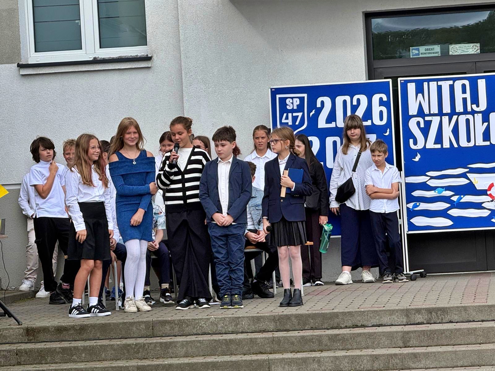 Grupa uczniów na szkolnych schodach, w tym dziewczynka w sweterku w paski śpiewająca do mikrofonu podczas występu artystycznego na rozpoczęcie roku szkolnego.