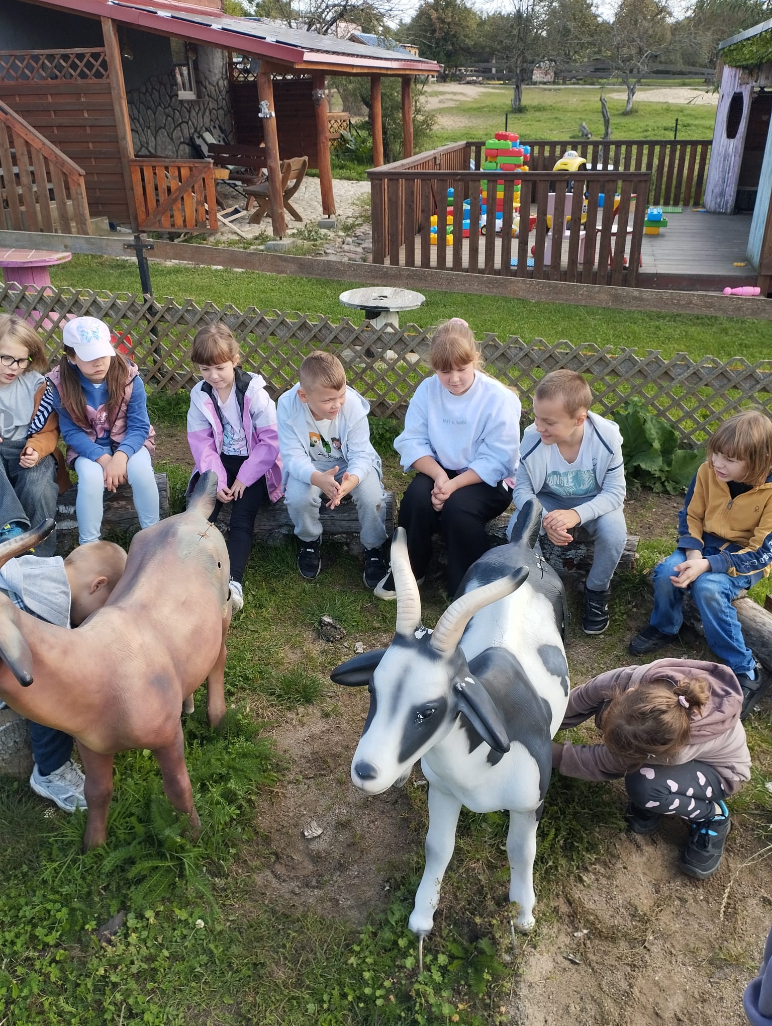 Dzieci siedzą na drewnianych ławkach na świeżym powietrzu, obok dużych figur zwierząt gospodarskich.