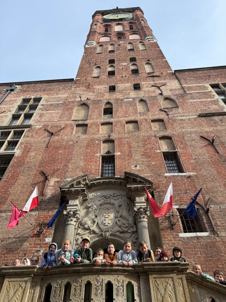Ceglana, zabytkowa wieża z dużym zegarem i ozdobnym, kamiennym portalem wejściowym. Na balkonie nad wejściem stoi grupa uczniów kl. 4b, a po bokach powiewają polskie i europejskie flagi. W tle widoczne jest błękitne niebo.