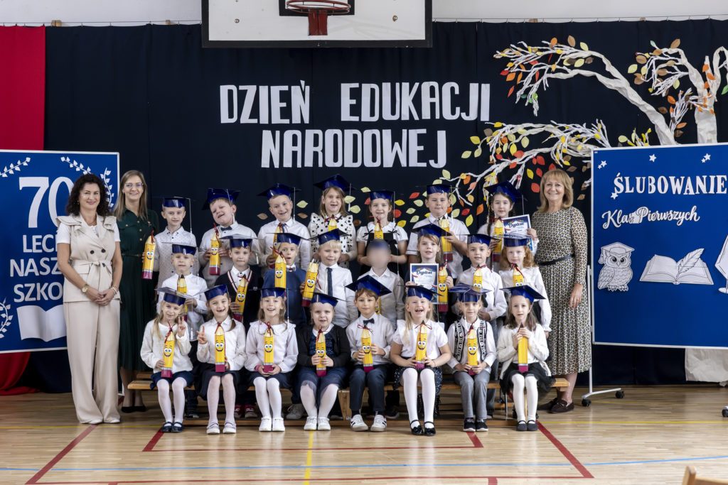 Uczniowie klasy pierwszej w biretach i strojach galowych pozują do pamiątkowego zdjęcia z wychowawczyniami po uroczystości pasowania na ucznia. W tle widoczne są dekoracje z okazji Dnia Edukacji Narodowej, jubileuszu 70-lecia szkoły oraz ślubowania pierwszoklasistów.