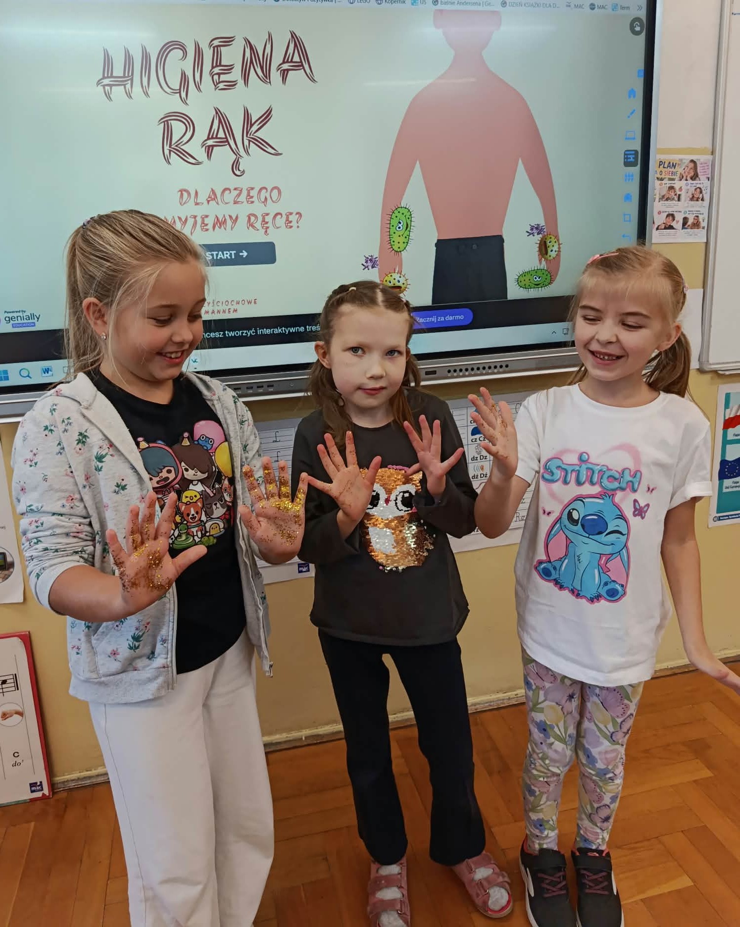 Trzy uczennice pokazują dłonie ozdobione kolorowym brokatem podczas zajęć o higienie rąk. Za nimi znajduje się prezentacja multimedialna dotycząca higieny dłoni.