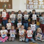 Grupa dzieci w klasie szkolnej prezentuje kolorowe prace plastyczne związane z higieną i zdrowiem. Uczniowie z dumą pokazują swoje ilustracje.