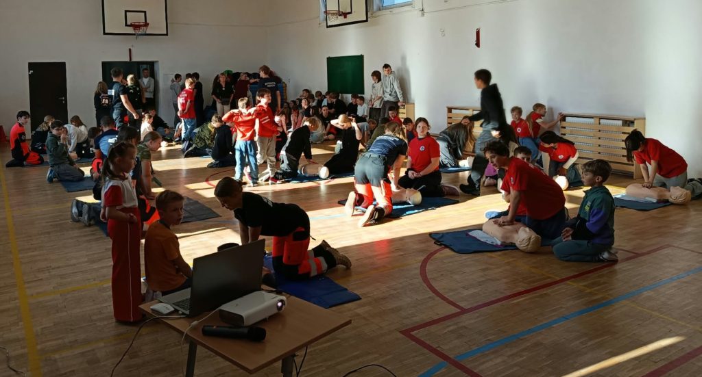 W sali gimnastycznej na podłodze leżą 3 rzędy fantomów przy nich klęczą po 2 lub 3 osoby. Jedna z osób wykonuje na fantomie masaż serca. Na pierwszym planie stolik z laptopem i rzutnikiem. W tle wyjście z sali.