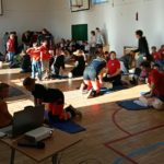 W sali gimnastycznej na podłodze leżą 3 rzędy fantomów przy nich klęczą po 2 lub 3 osoby. Jedna z osób wykonuje na fantomie masaż serca. Na pierwszym planie stolik z laptopem i rzutnikiem. W tle wyjście z sali.