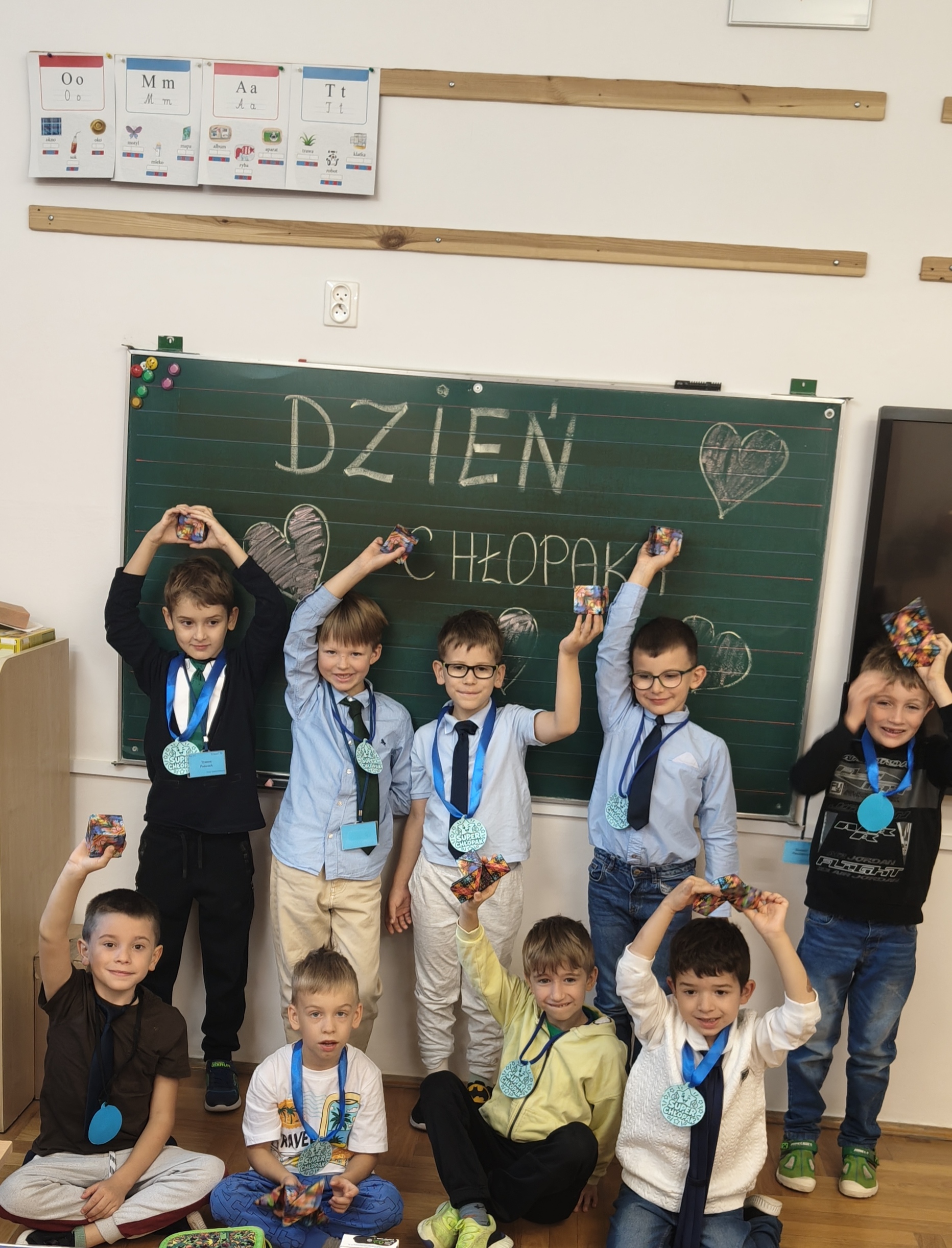 Grupa dziesięciu chłopców w klasie pozuje pod zieloną tablicą z napisem „Dzień Chłopaka”, trzymając w rękach małe paczki z prezentami i medale na szyjach