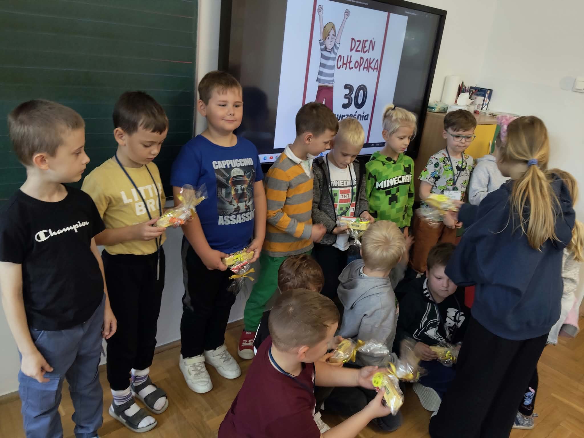 Chłopcy stojący w klasie obok tablicy interaktywnej z napisem „Dzień Chłopaka”, z uwagą oglądający upominki wręczane przez koleżankę.