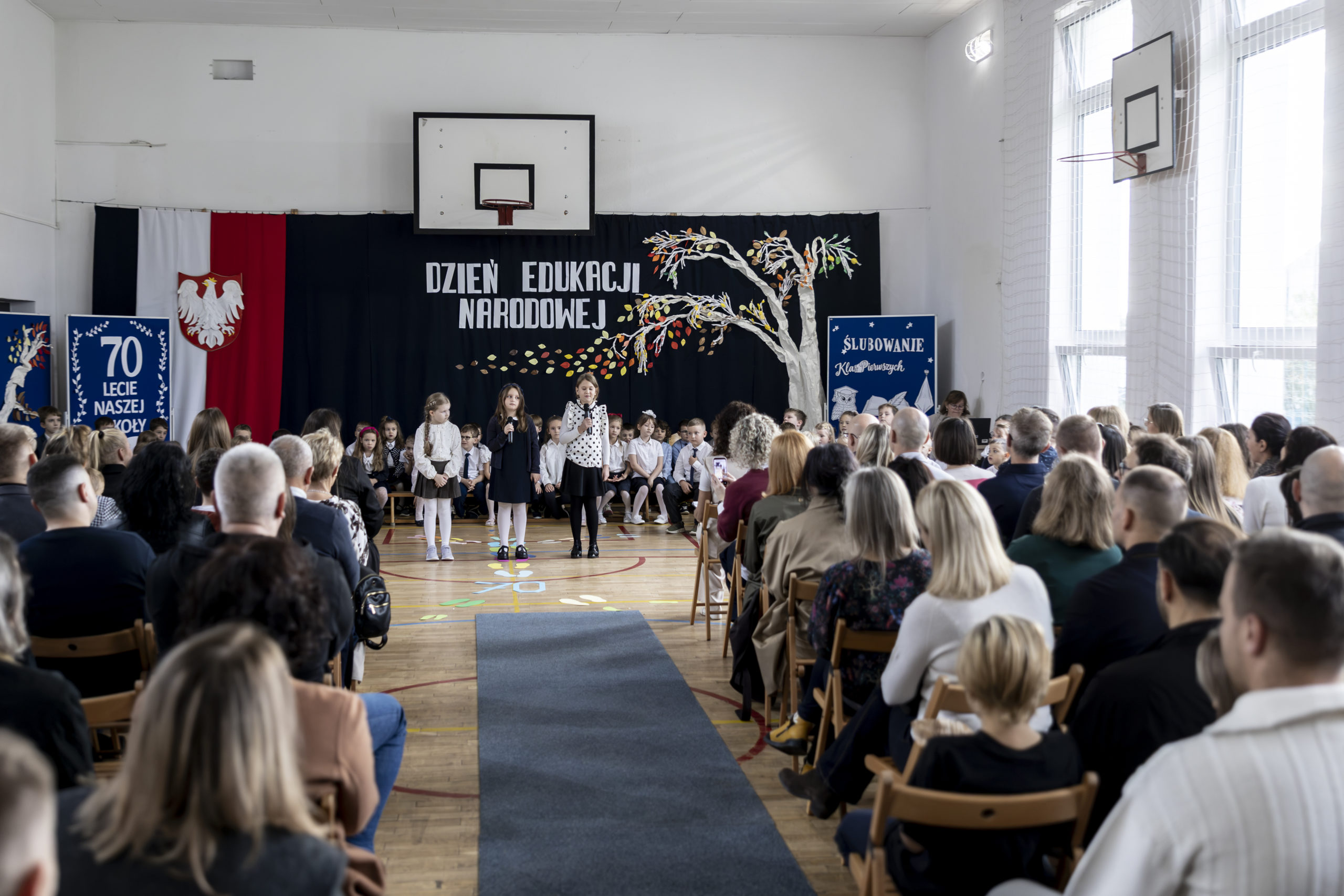 Uczniowie klas pierwszych prezentują przygotowany program artystyczny podczas uroczystości ślubowania, a zgromadzeni rodzice i goście oglądają występ. W tle widoczne są dekoracje z okazji Dnia Edukacji Narodowej, ślubowania pierwszoklasistów oraz 70-lecia szkoły.