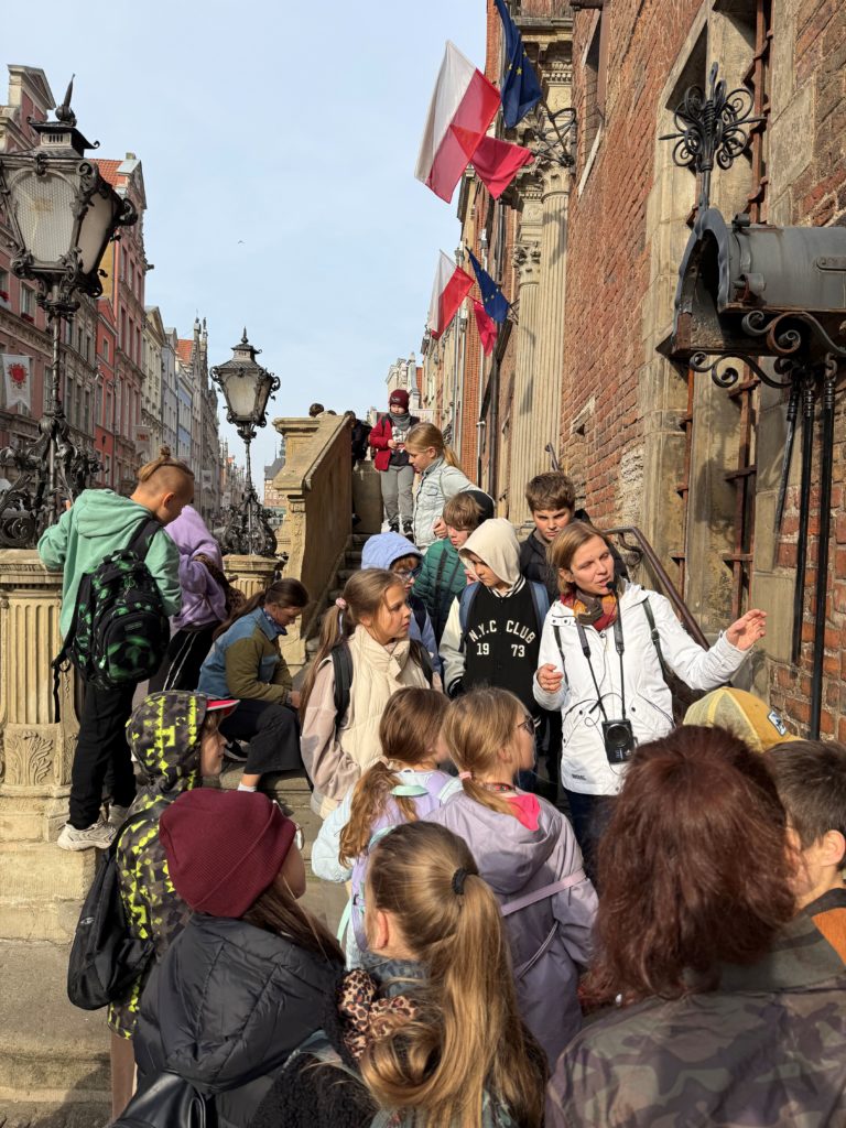 Grupa dzieci i opiekunów stoi na kamiennych schodach przy zabytkowej, ceglanej kamienicy. Na elewacji wiszą polskie i unijne flagi, a wzdłuż ulicy znajdują się ozdobne, historyczne latarnie. W tle widać zabytkową strarówkę Gdańska.