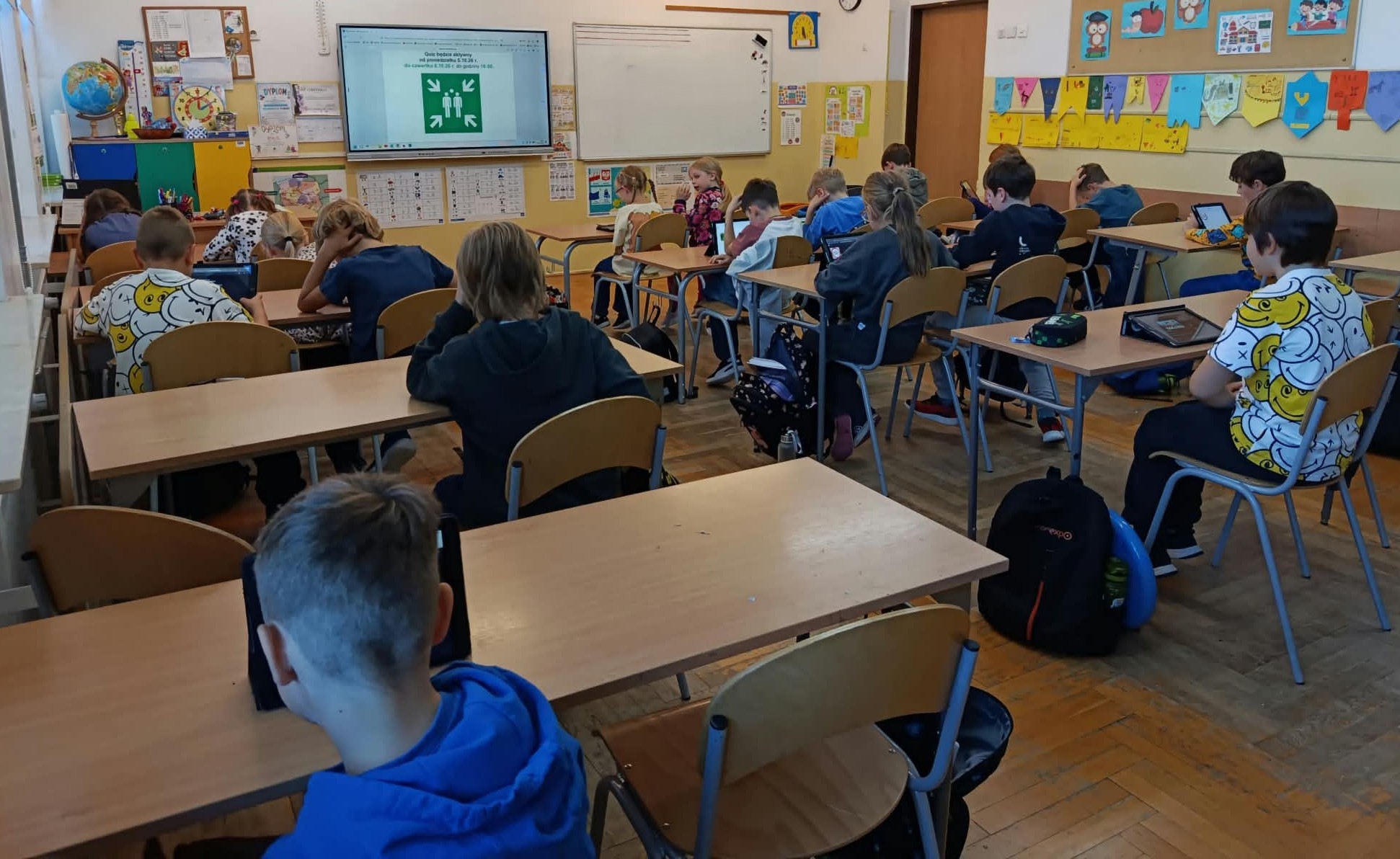 W sali lekcyjnej w trzech rzędach ławek siedzą obróceni tyłem uczniowie klasy 2c. Przed uczniami leżą na ławce IPady, na którym wyświetlają się pytania quizu. W tle tablica multimedialna z plakatem konkursu „Bezpieczeństwo w szkole i na drodze” oraz znakiem ewakuacyjnym.