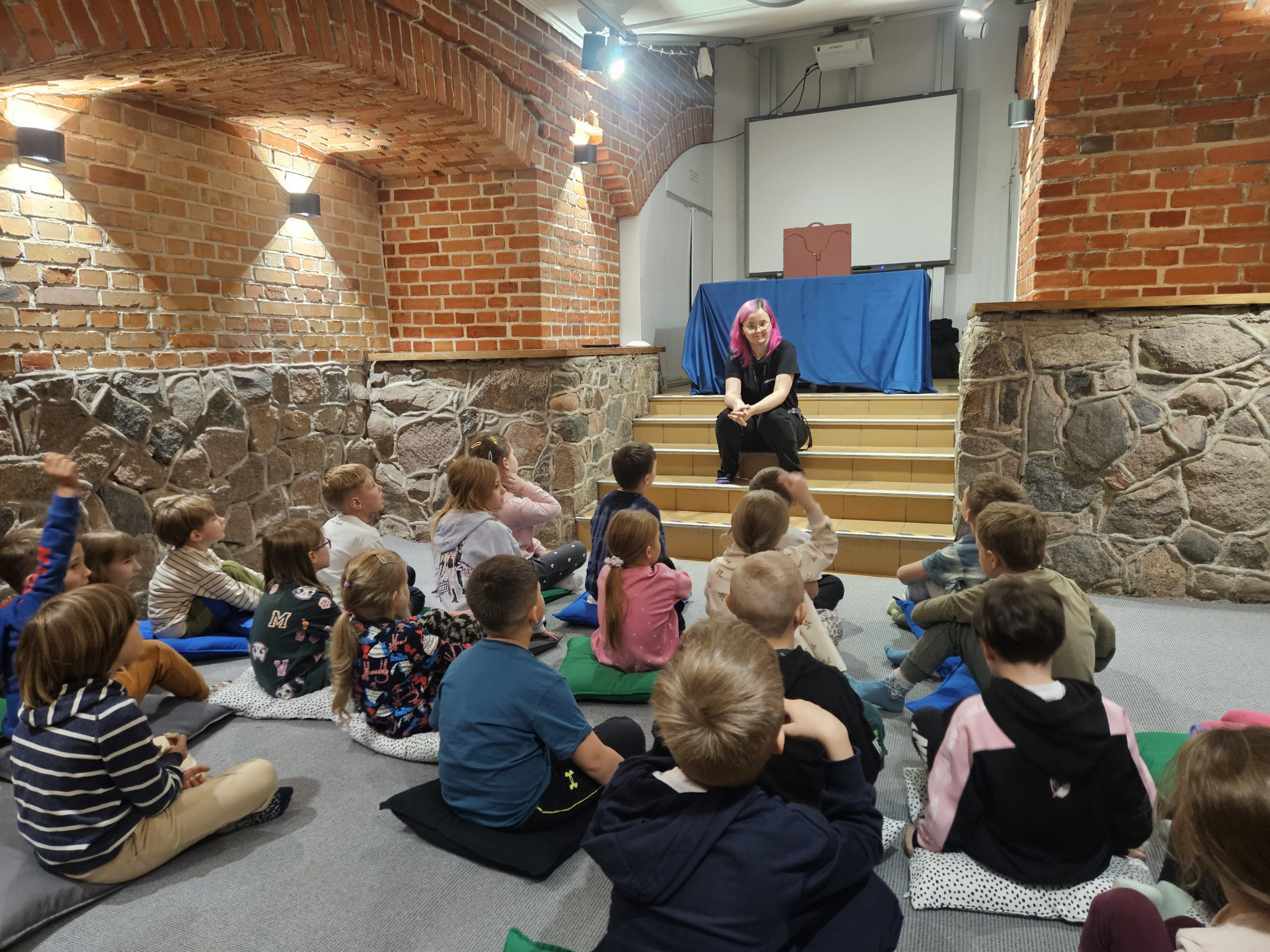 Uczniowie klasy 1A siedzą na poduszkach i słuchają prowadzącej podczas warsztatów edukacyjnych w Hevelianum. Zdjęcie nr 2 Uczniowie klasy 1A poznają zasady działania energii elektrycznej, korzystając z interaktywnej instalacji edukacyjnej w Hevelianum.