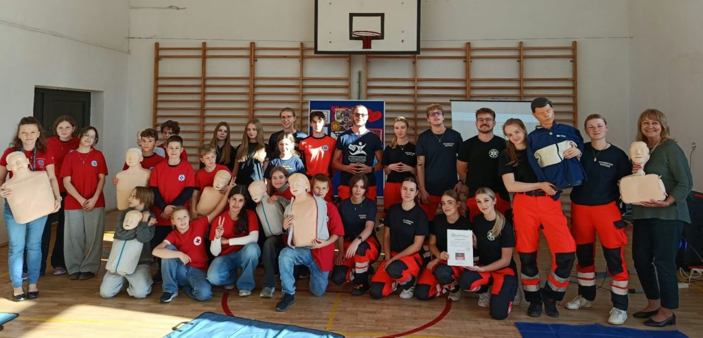 W sali gimnastycznej na tle drabinek i tablicy z plakatem stoją 2 rzędy ludzi oraz 1 rząd przed nimi kuca. Ponad trzydziestoosobowa grupa ludzi pozuje do zdjęcia, część z nich z fantomami w rękach.