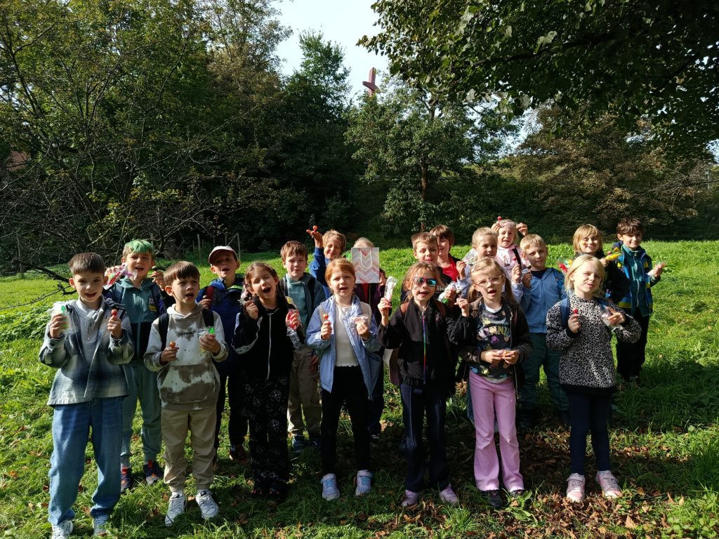 Duża grupa dzieci stojąca na trawie pod drzewami, trzymająca w dłoniach drobne upominki.