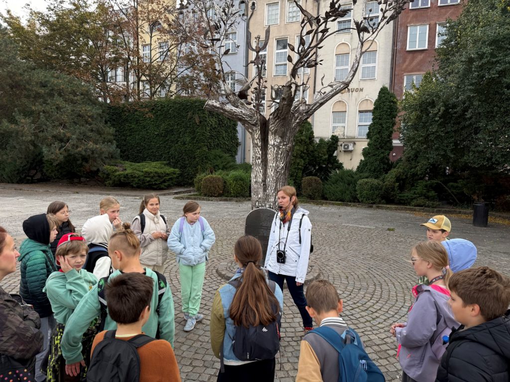 Grupa dzieci podczas wycieczki szkolnej słucha przewodniczki opowiadającej o metalowej rzeź-bie drzewa na brukowanym placu w otoczeniu zabytkowych kamienic i zieleni.