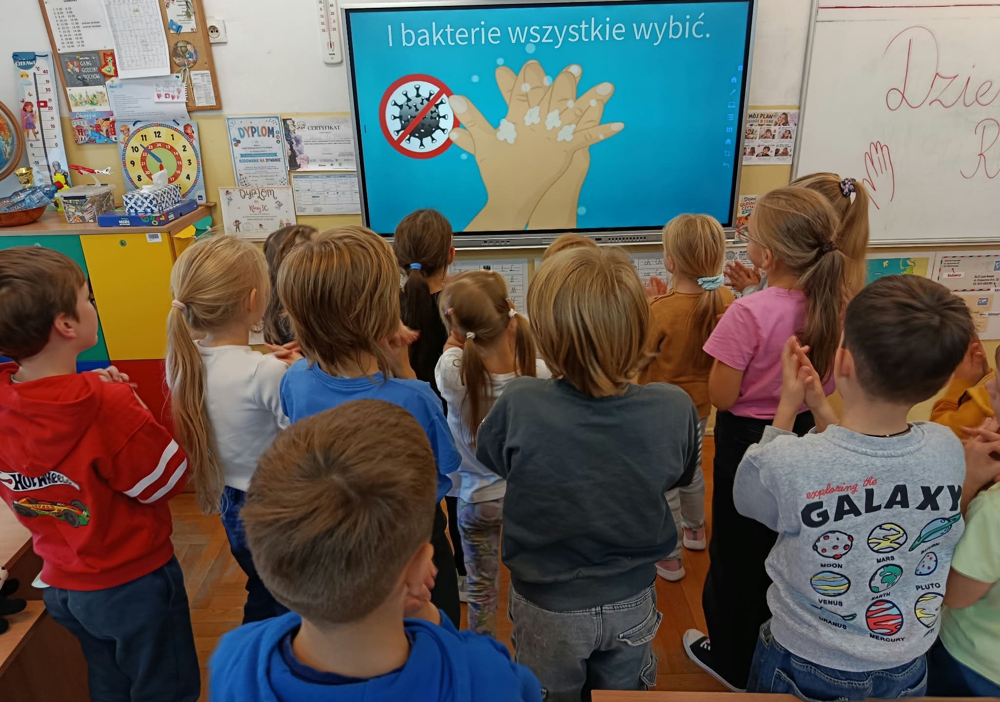Dzieci stoją przed tablicą interaktywną, na której wyświetlono ilustrację dłoni pokrytych mydłem oraz hasło zachęcające do usuwania bakterii podczas mycia rąk.