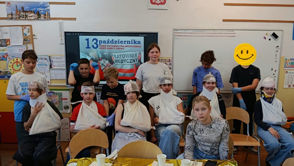 Na tle tablicy z napisem 13 października Dzień Ratownictwa Medycznego ekipa ratunkowa zakładająca opatrunki i temblaki pozorantom.