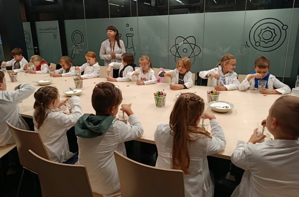 Dookoła długiego i jasnego stołu ustawionego w sali warsztatowej Muzeum Bursztynu siedzą na krzesłach uczniowie klasy 2c. Każdy z uczniów miesza łyżką w plastikowym kubeczku napełnionym wodą z solą i odławia bursztyn. Obok stoi kustoszka muzeum.