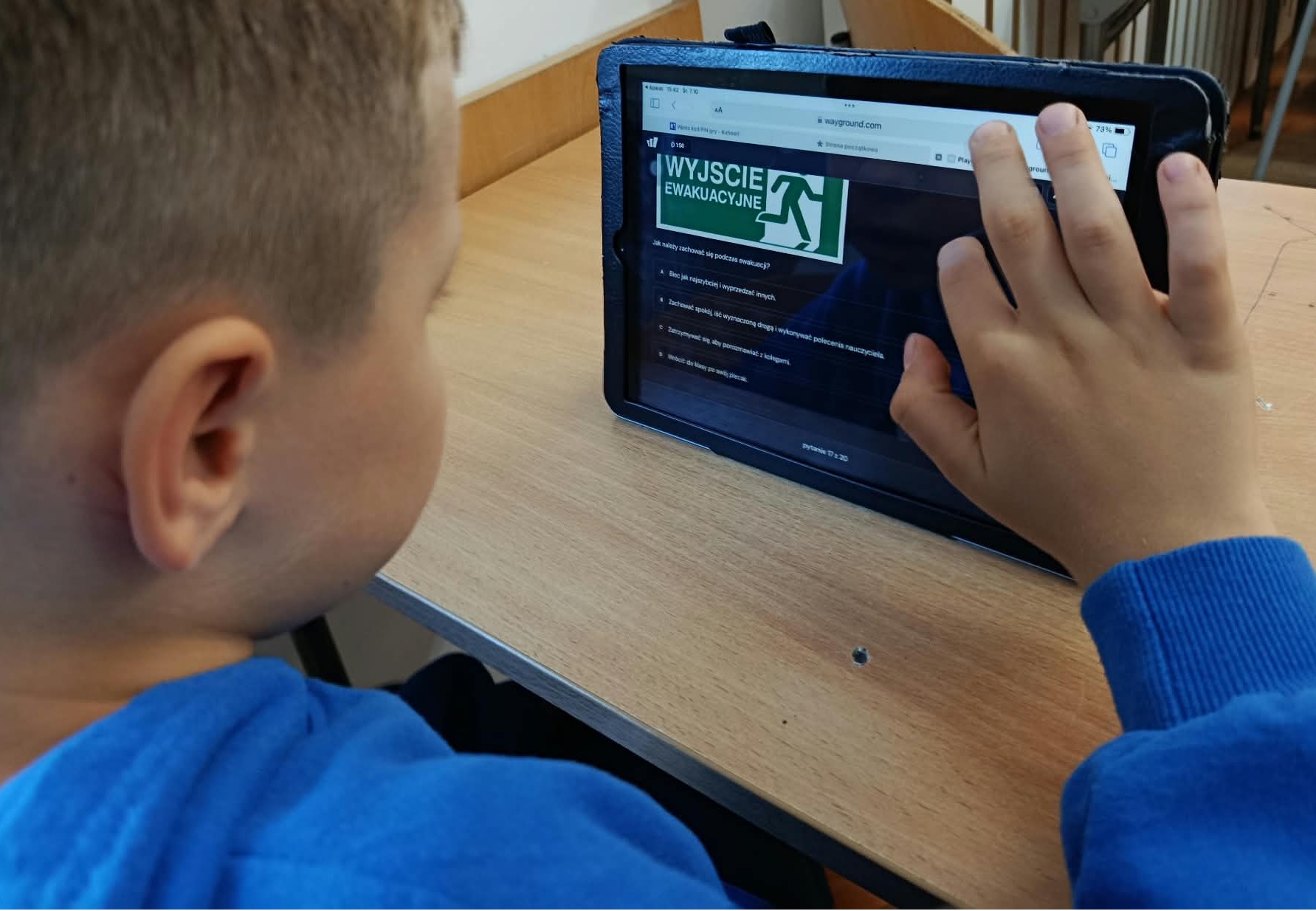 W sali lekcyjnej w ławce siedzi uczeń klasy 2c. Przed nim stoi IPad wyświetlający pytanie z Quizu i znak ewakuacyjny.