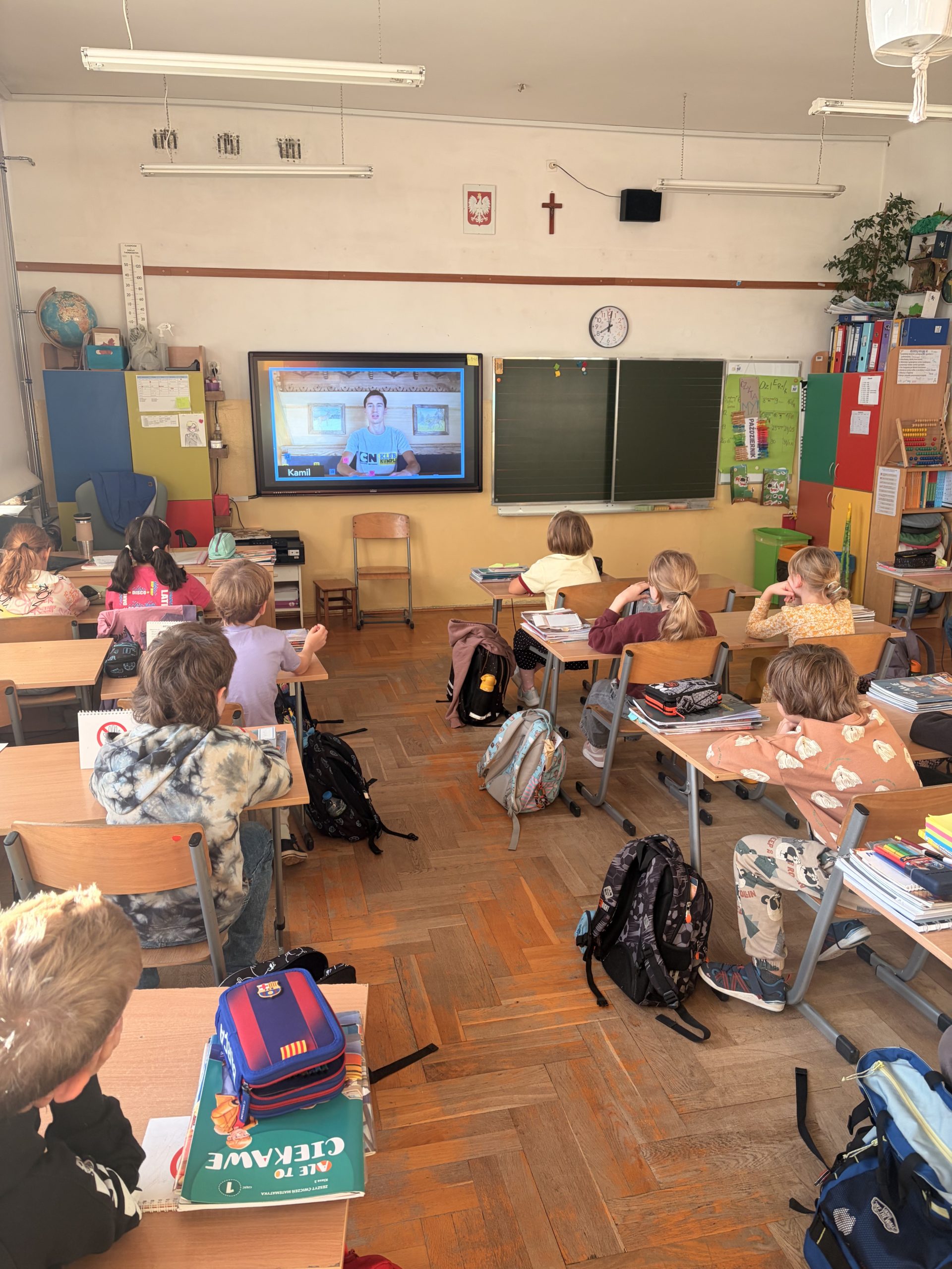 Widok na klasę szkolną z perspektywy tyłu sali. Dzieci siedzą przy dwuosobowych ławkach i uważnie patrzą na ekran interaktywny, na którym włączony jest film z prezentacją.