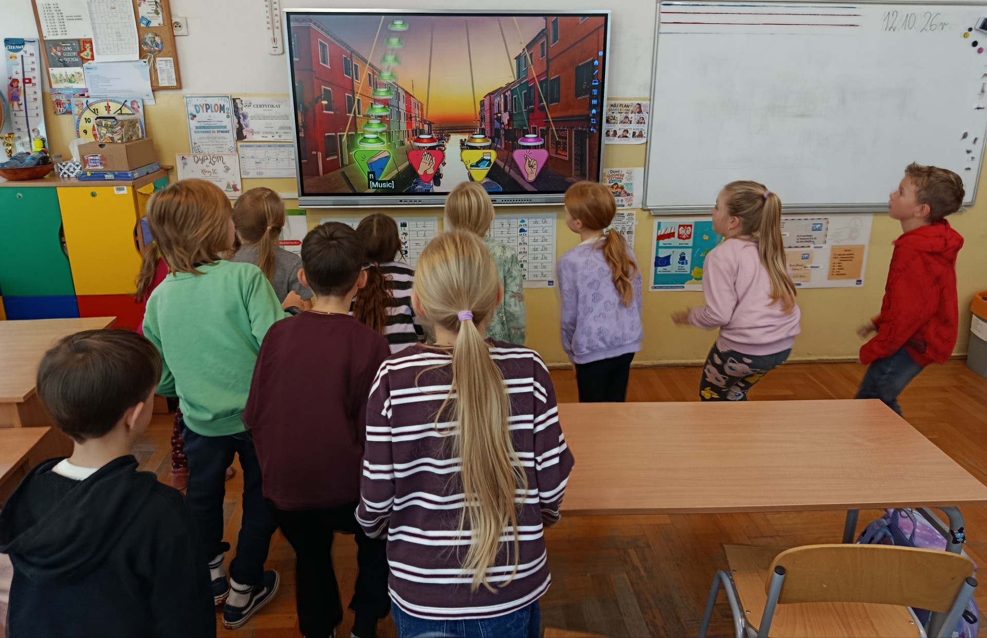  Dziesięciu uczniów klasy 2c wyklaskujących rytm stoi obróconych twarzą do tablicy multimedialnej.