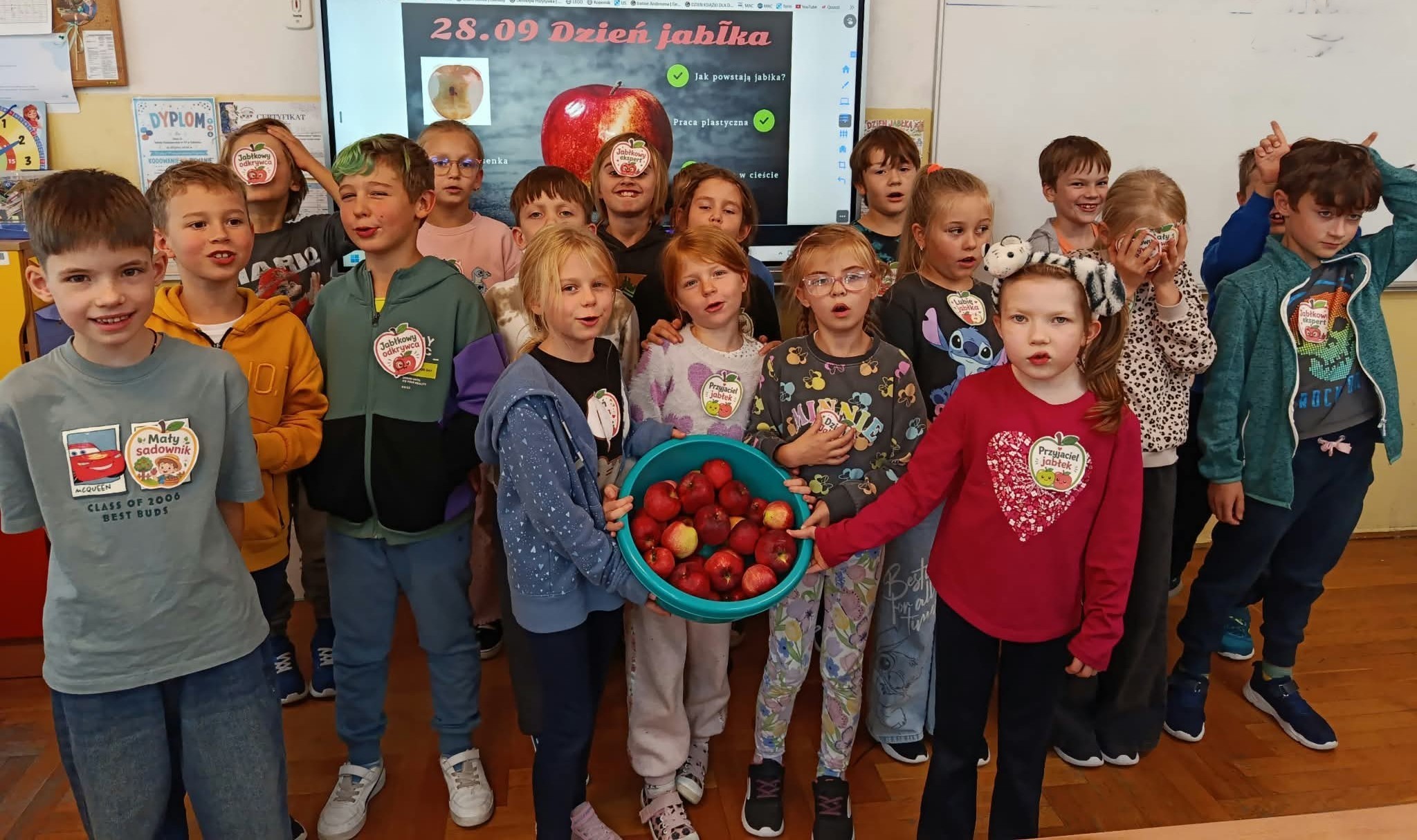 Na tle tablicy multimedialnej wyświetlającej zadania z okazji Dnia Jabłka stoi grupa uczniów klasy 2c z odznakami sadownika przypiętymi do bluzeczki. Na pierwszym planie dziewczynki trzymające miskę z jabłkami.