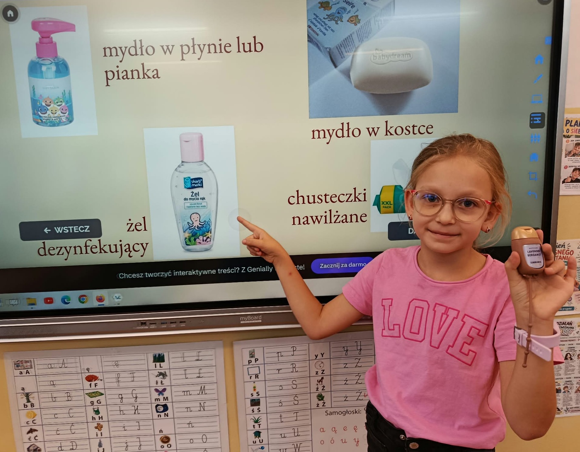 Uczennica w różowej koszulce wskazuje na prezentację multimedialną przedstawiającą różne rodzaje środków do higieny rąk, m.in. mydło w płynie, mydło w kostce i żel dezynfekujący.