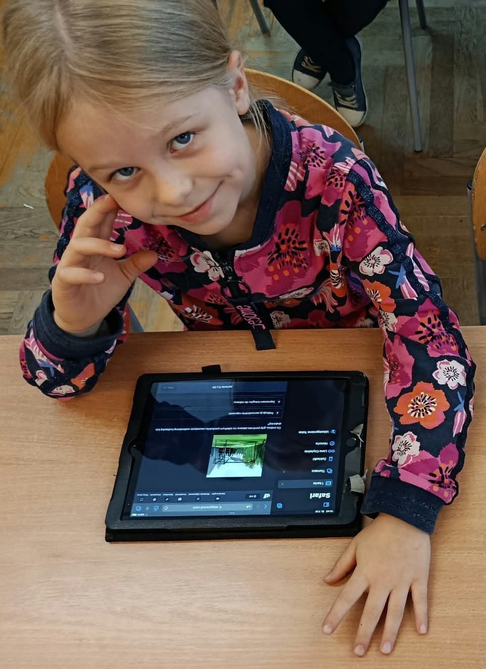 Na krzesełku siedzi uśmiechająca się uczennica klasy 2c. Przed nią na ławce leży IPad wyświetlający pytanie z Quizu.