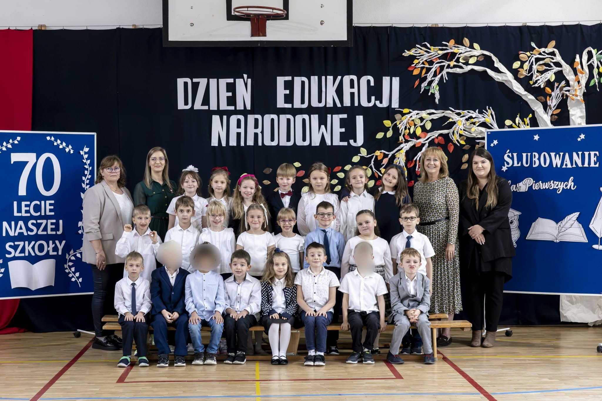 Uczniowie klasy pierwszej w strojach galowych pozują do pamiątkowego zdjęcia z wychowawczyniami i dyrekcją po uroczystości pasowania na ucznia. W tle widoczne są dekoracje z okazji Dnia Edukacji Narodowej, jubileuszu 70-lecia szkoły oraz ślubowania pierwszoklasistów.