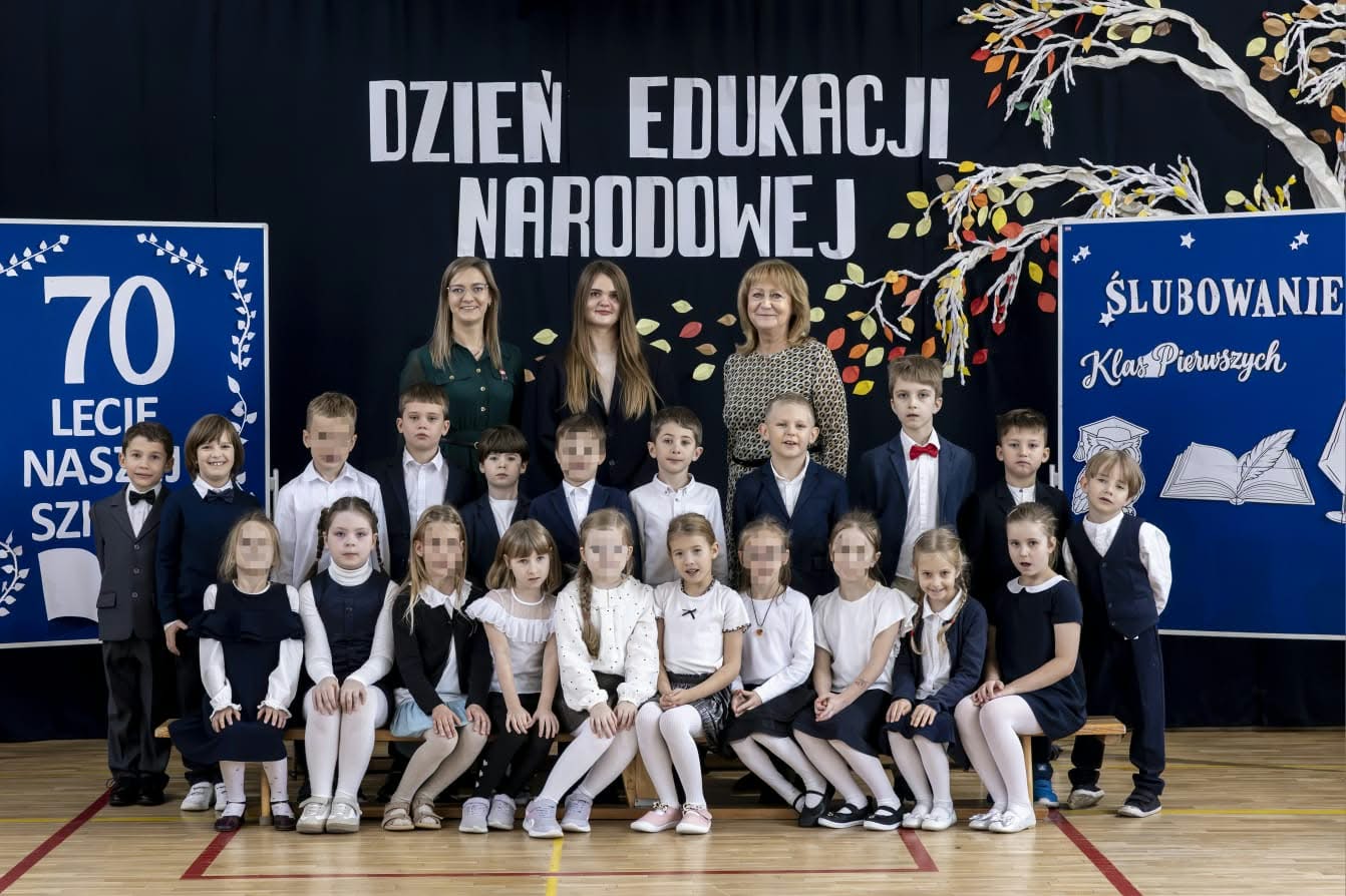 Uczniowie klasy pierwszej w strojach galowych pozują do pamiątkowego zdjęcia z wychowawczyniami i dyrekcją po uroczystości pasowania na ucznia. W tle widoczne są dekoracje z okazji Dnia Edukacji Narodowej, jubileuszu 70-lecia szkoły oraz ślubowania pierwszoklasistów.