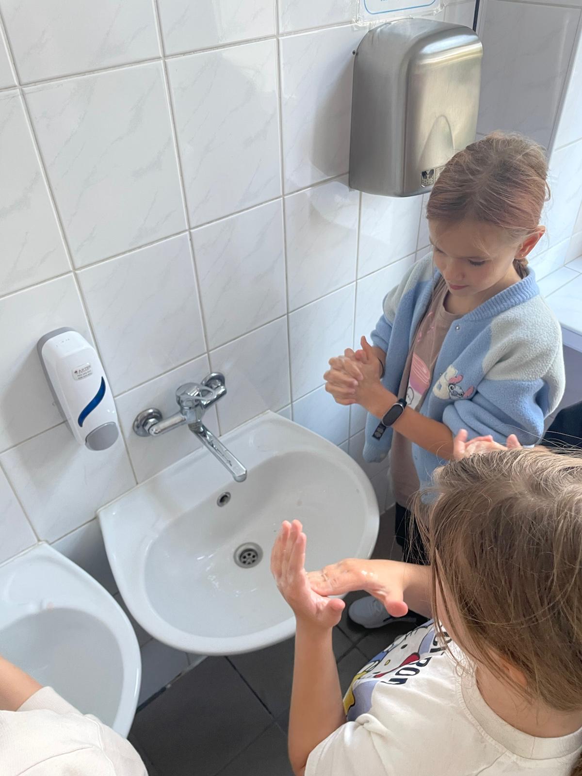 Uczennice ćwiczą prawidłowe mycie rąk przy szkolnych umywalkach. Jedna dziewczynka pociera dłonie, a druga rozprowadza mydło na dłoni, ucząc się zasad higieny.