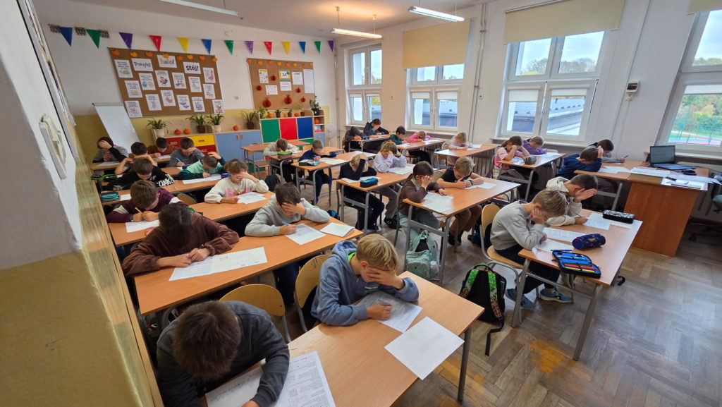 W szkolnej sali siedzą uczniowie pochyleni i zamyśleni nad kartkami konkursowymi.