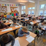 W szkolnej sali siedzą uczniowie pochyleni i zamyśleni nad kartkami konkursowymi.