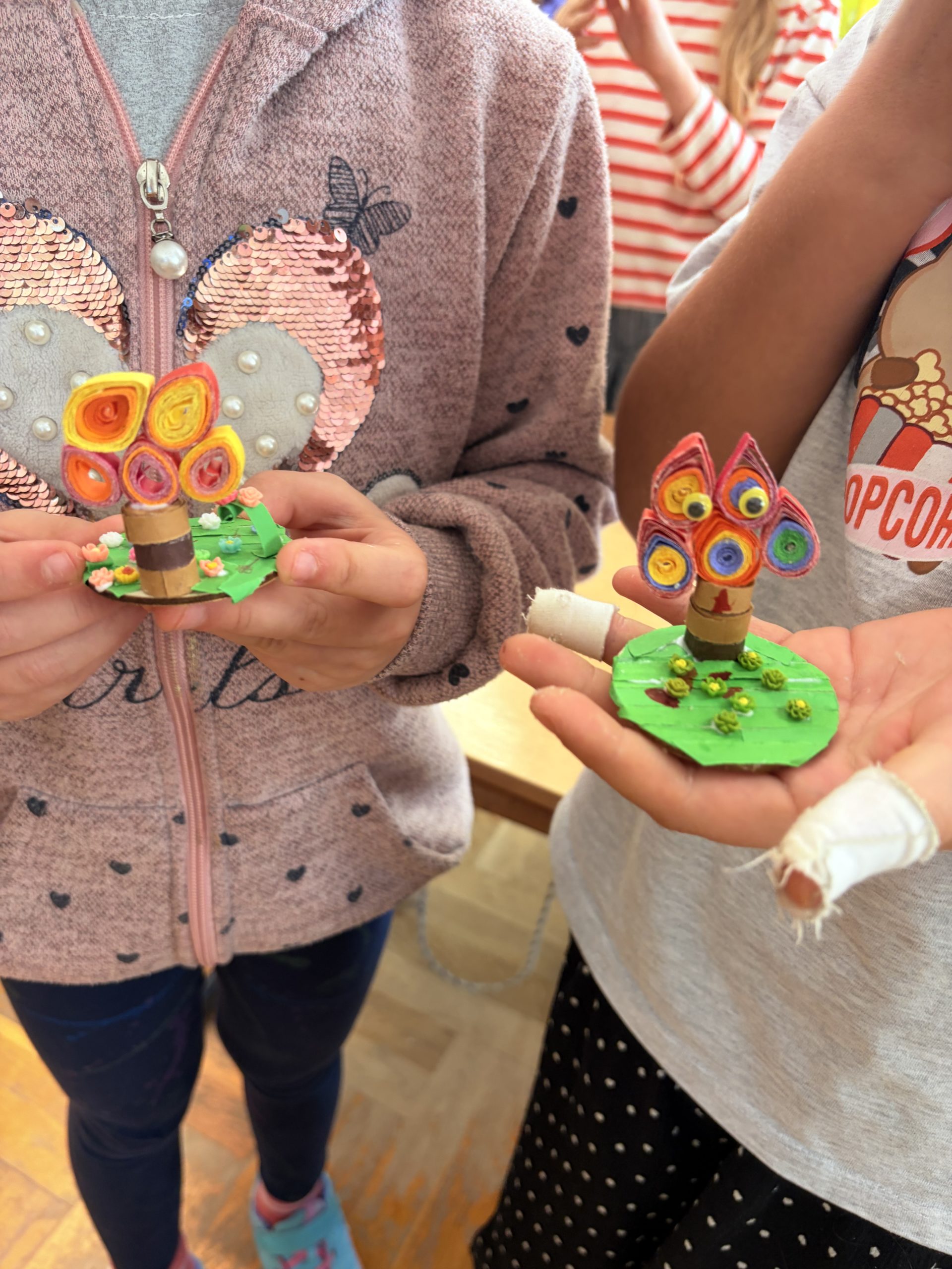 Dłonie uczniów trzymające dwa miniaturowe drzewka z quillingu na szkolnej ławce. Drzewka mają brązowe pnie i barwne korony ze zwiniętych pasków papieru, jedno stoi na kwadratowej, a drugie na okrągłej zielonej podstawce.