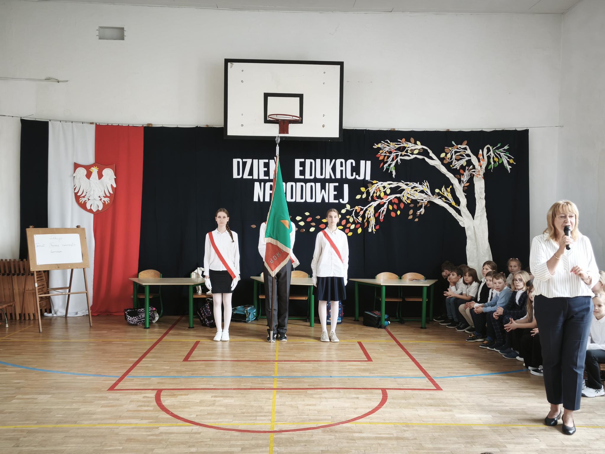  Uczniowie w strojach galowych, z biało-czerwonymi szarfami, stoją przy poczcie sztandarowym podczas uroczystości z okazji Dnia Edukacji Narodowej. Obok nauczycielka przemawia do zgromadzonych uczniów.