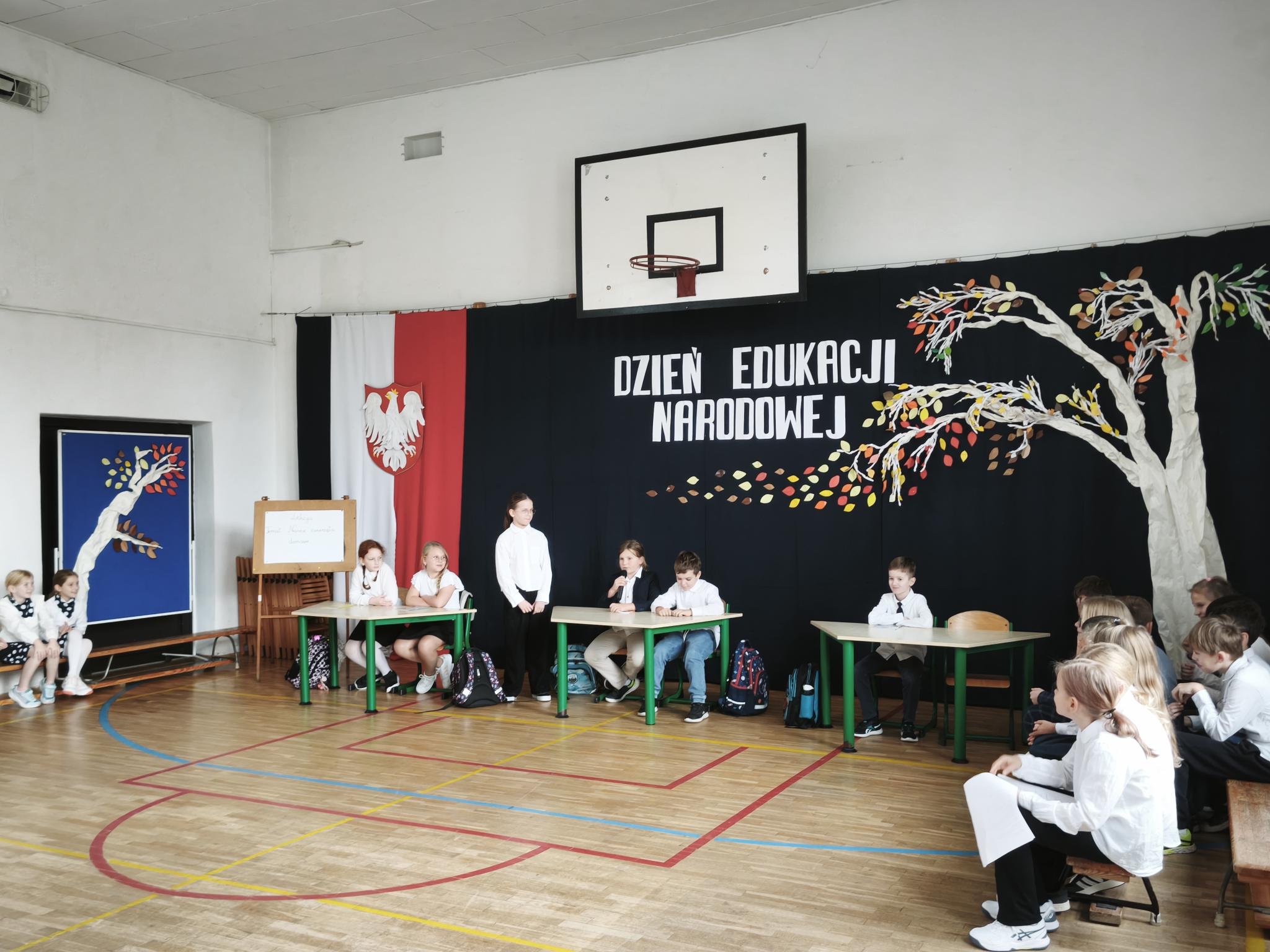 Uczniowie w strojach galowych uczestniczą w inscenizacji z okazji Dnia Edukacji Narodowej, siedząc przy szkolnych stolikach na tle uroczystej dekoracji.