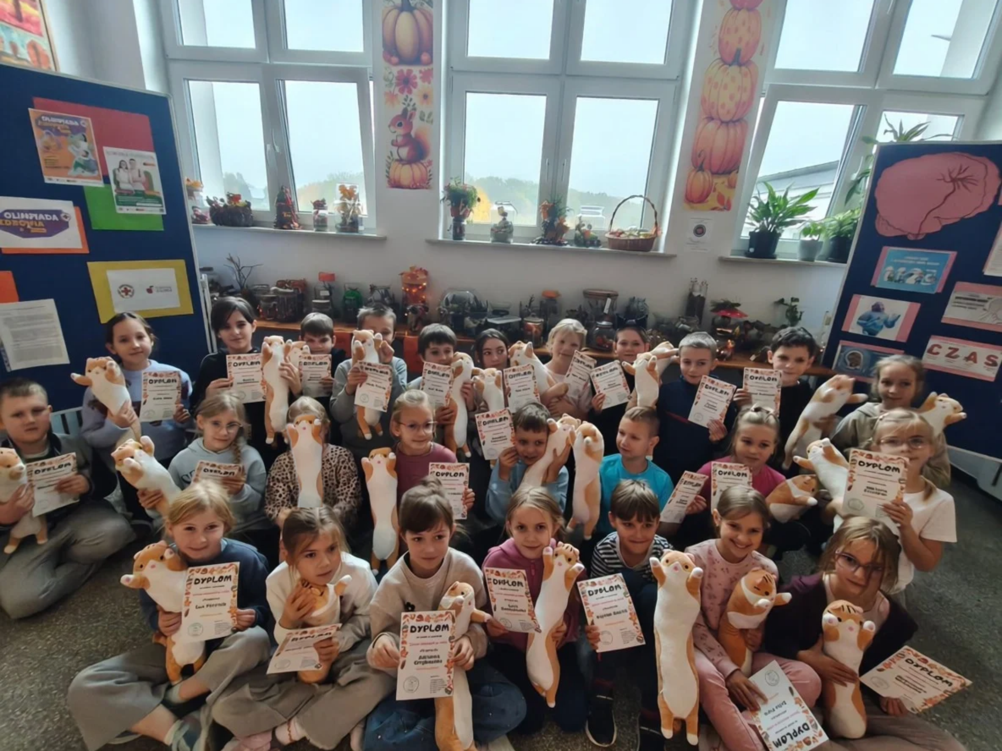 Grupa uczniów pozuje do wspólnego zdjęcia, trzymając pluszowe maskotki i dyplomy za udział w konkursie. Dzieci siedzą  w kilku rzędach na korytarzu, w tle widać okna, jesienne dekoracje oraz tablice informacyjne.