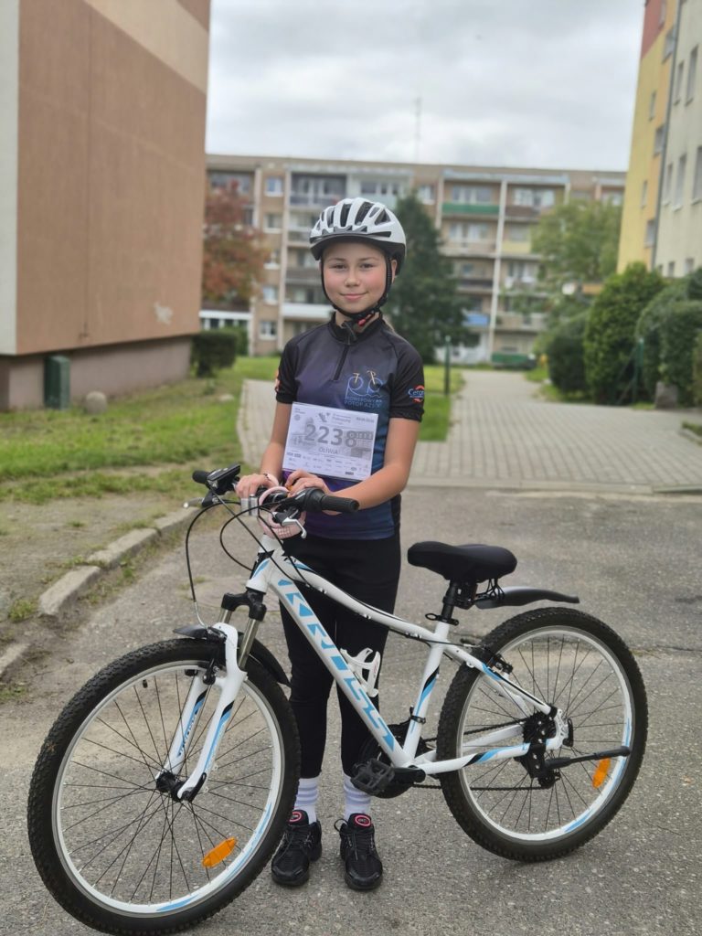 Dziewczyna w kasku rowerowym stoi z rowerem na chodniku. Na koszulce ma przyczepiony numer startowy biegu charytatywnego. Jest uśmiechnięta.