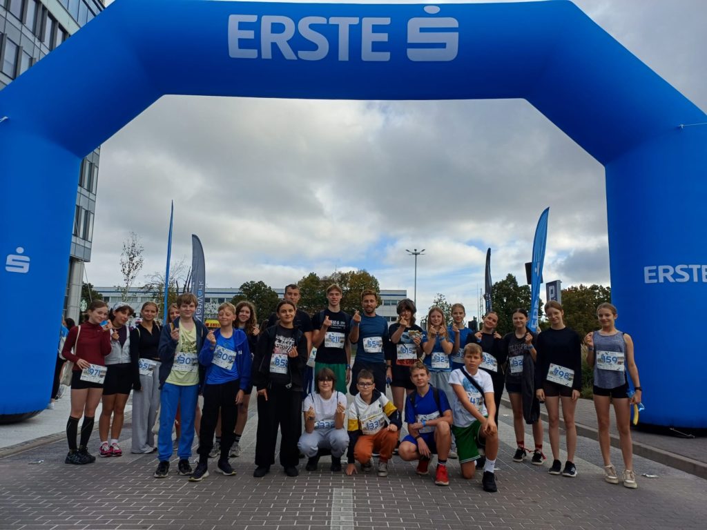 Grupa uczniów i opiekun pozuje do zdjęcia pod wielką bramą startową biegu charytatywnego. Wszyscy mają numery startowe i uśmiechają się do aparatu. Pokazują do aparatu finger hearts (serduszka z palców).