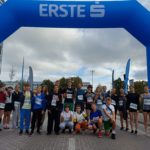 Grupa uczniów i opiekun pozuje do zdjęcia pod wielką bramą startową biegu charytatywnego. Wszyscy mają numery startowe i uśmiechają się do aparatu. Pokazują do aparatu finger hearts (serduszka z palców).