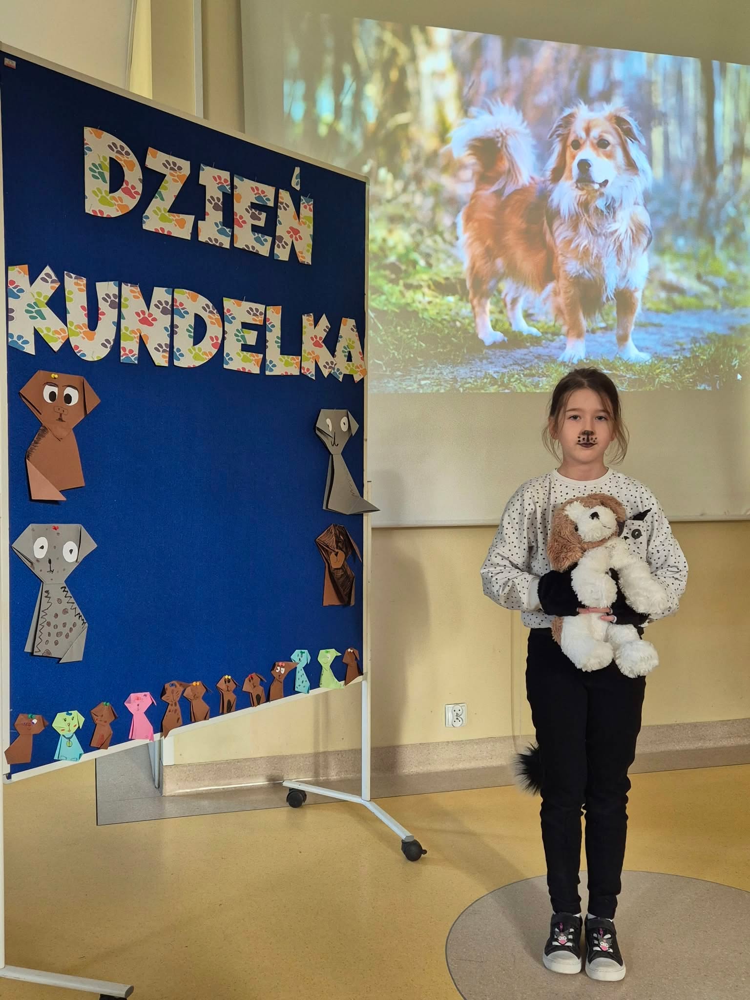 Dziewczynka stoi na tle tablicy z napisem „Dzień Kundelka”. Na twarzy ma namalowany czarny nosek pieska, a w rękach trzyma pluszową maskotkę przedstawiającą psa. W tle widoczny jest ekran z rzutnika wyświetlający zdjęcie kudłatego kundelka.