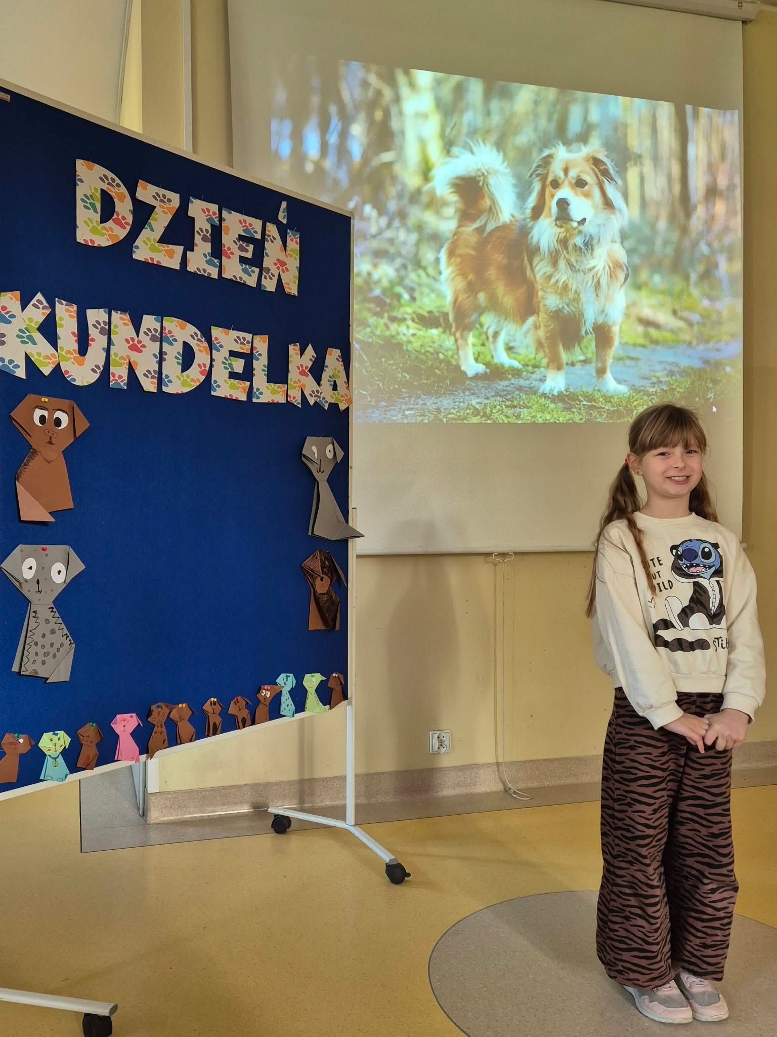 Dziewczynka uśmiecha się stojąc obok tablicy informacyjnej z napisem „Dzień Kundelka”. Na tablicy widoczne są papierowe origami przedstawiające psy. W tle za nią znajduje się rzutnik z wyświetlonym zdjęciem psa.