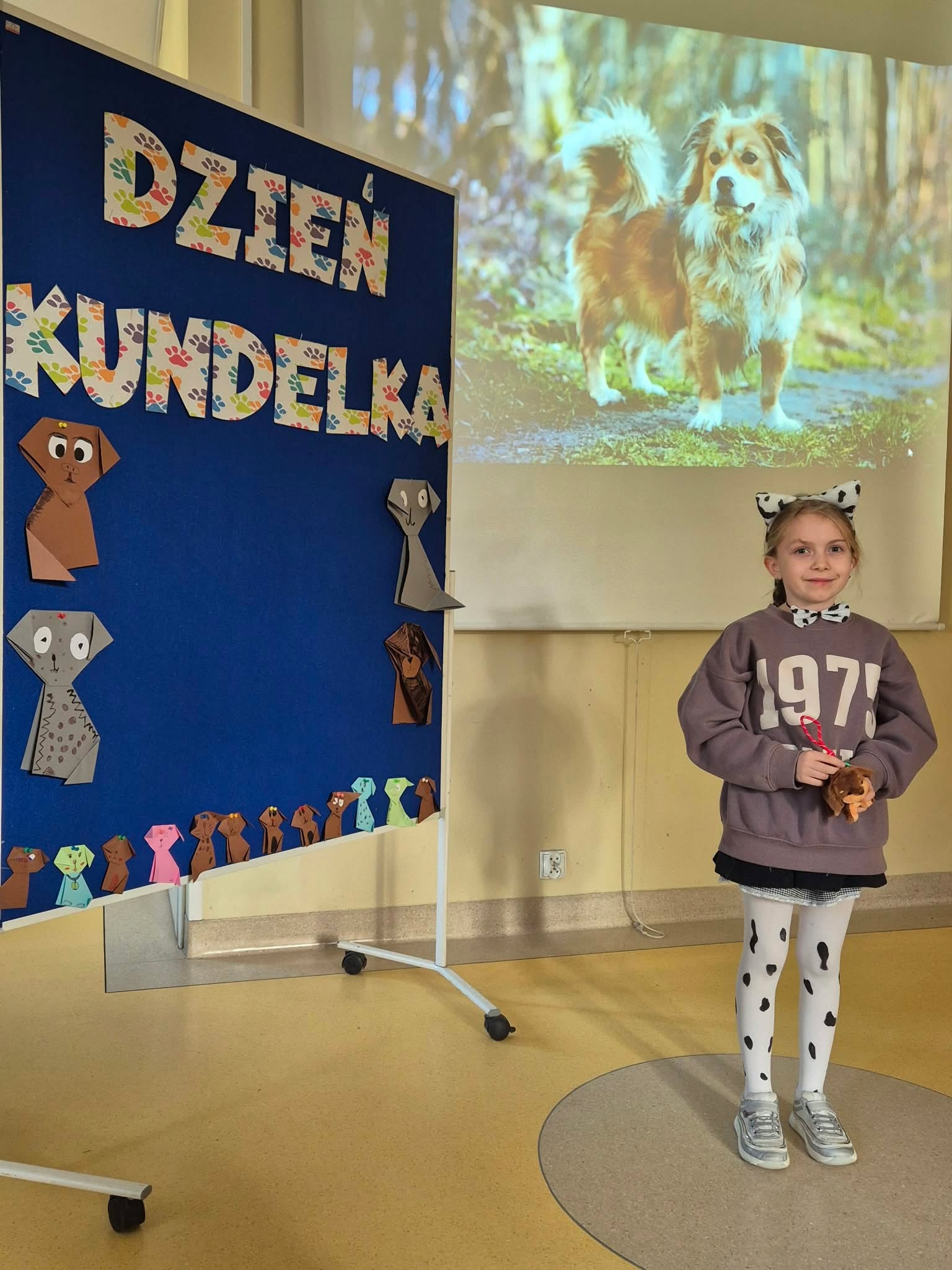 Dziewczynka uśmiecha się stojąc obok tablicy informacyjnej z napisem „Dzień Kundelka”. Na tablicy widoczne są papierowe origami przedstawiające psy. W tle za nią znajduje się rzutnik z wyświetlonym zdjęciem psa.