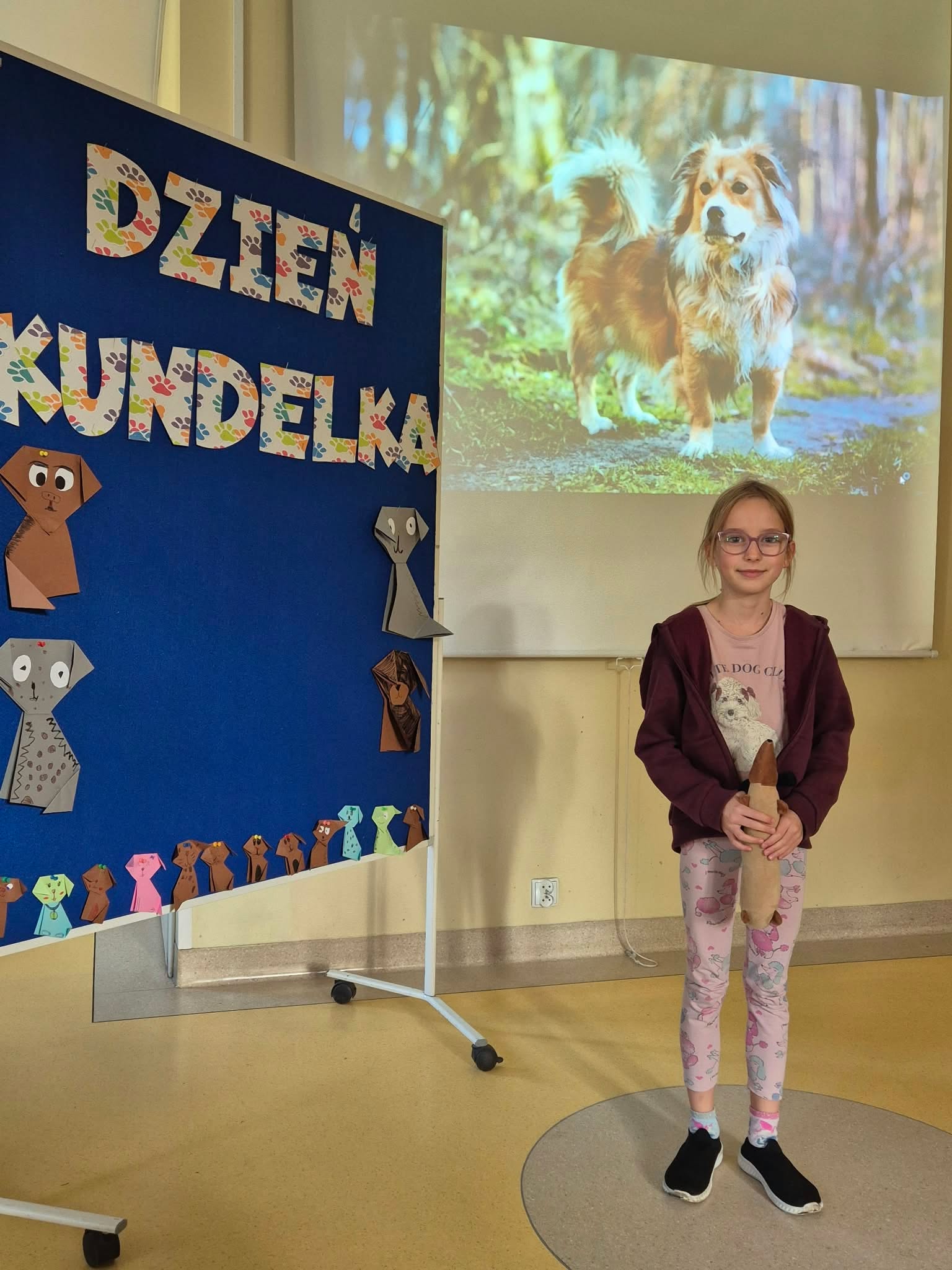 Dziewczynka uśmiecha się stojąc obok tablicy informacyjnej z napisem „Dzień Kundelka”. Na tablicy widoczne są papierowe origami przedstawiające psy. W tle za nią znajduje się rzutnik z wyświetlonym zdjęciem psa.