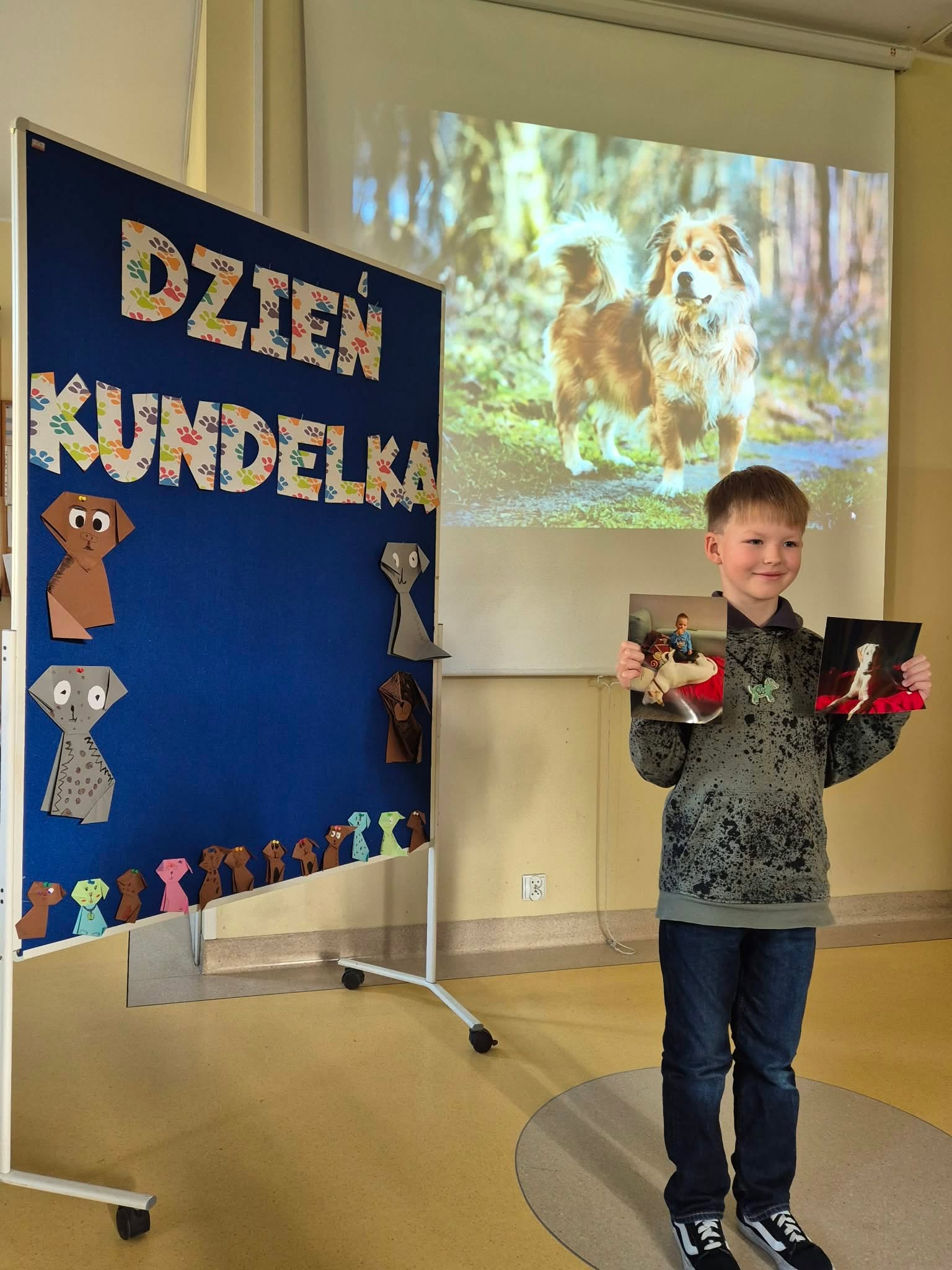 Chłopiec stoi na tle tablicy z napisem „Dzień Kundelka”. W obu dłoniach trzyma i prezentuje zdjęcia przedstawiające psa. Na ścianie za nim wyświetlany jest za pomocą rzutnika duży obraz puszystego czworonoga